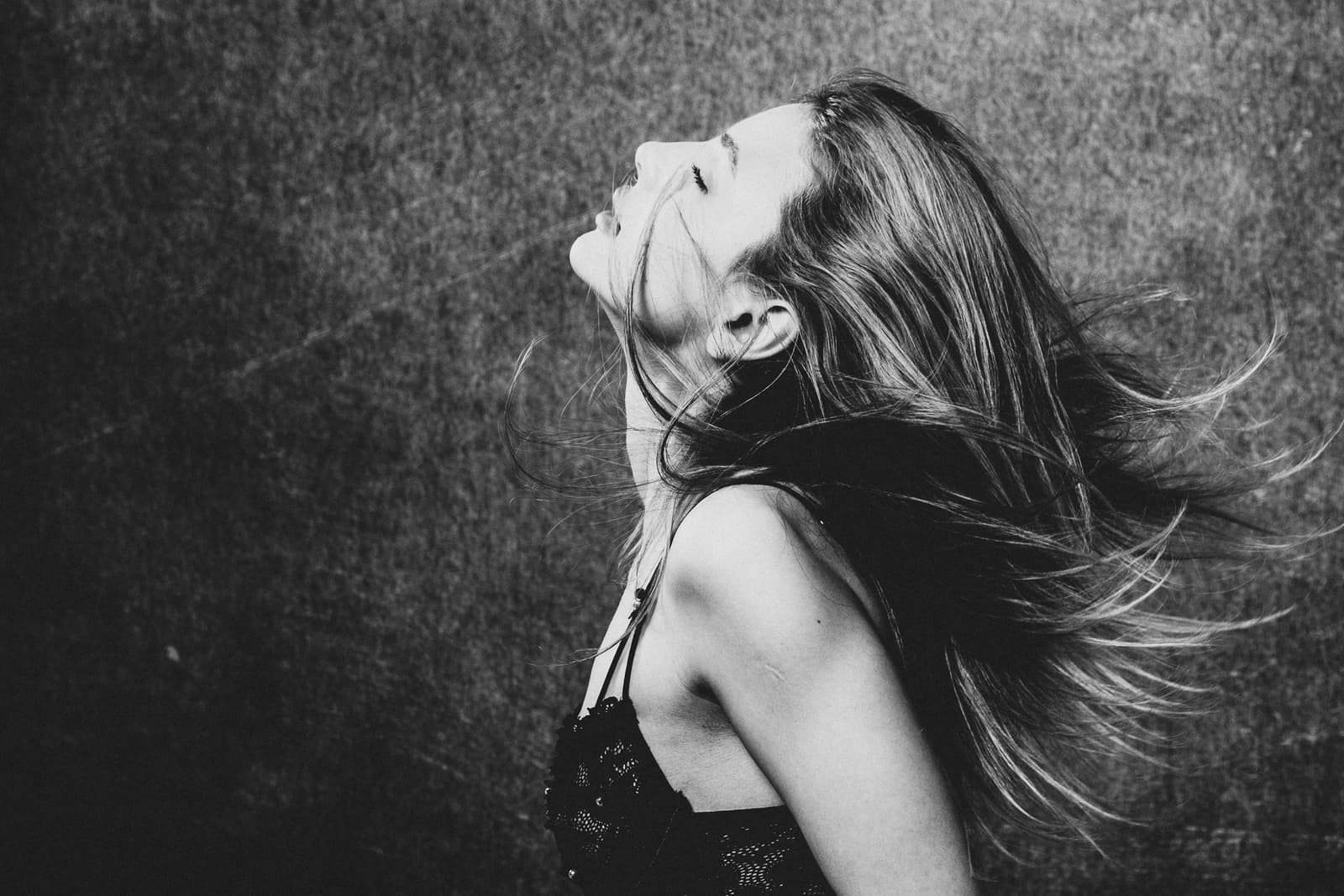 A black-and-white profile shot of a person with eyes closed and head tilted back. Their long hair is swept to one side, capturing movement. They are wearing a dark, textured top with thin straps against a mottled grey textured background.
