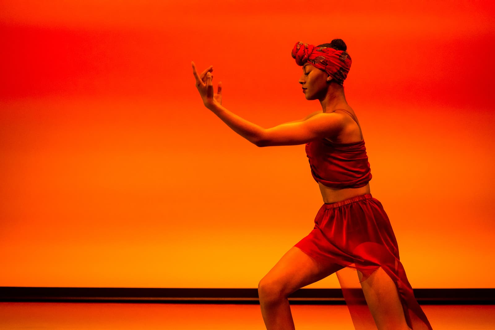 A profile view of a dancer in motion against a bright, solid orange-red background. The dancer wears a red headwrap, a red midriff-baring top, and a loose red skirt. One arm is extended upwards with fingers poised in a delicate gesture, while the body is angled in a wide stance.