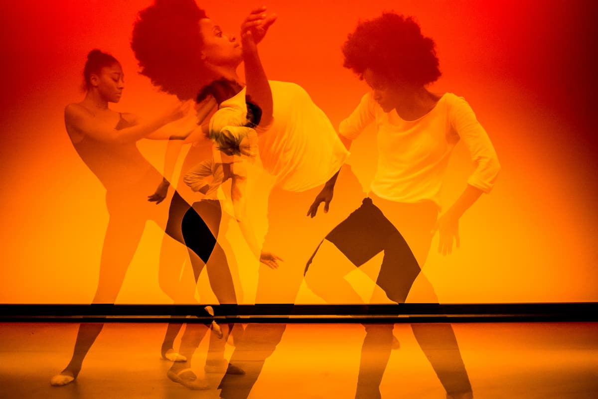 Multiple-exposure photograph of dancers in motion against a vibrant orange and yellow gradient background, creating overlapping translucent figures and limbs in various poses. The dancers wear simple athletic tops and shorts and are positioned near a horizontal black bar running across the lower third of the frame.