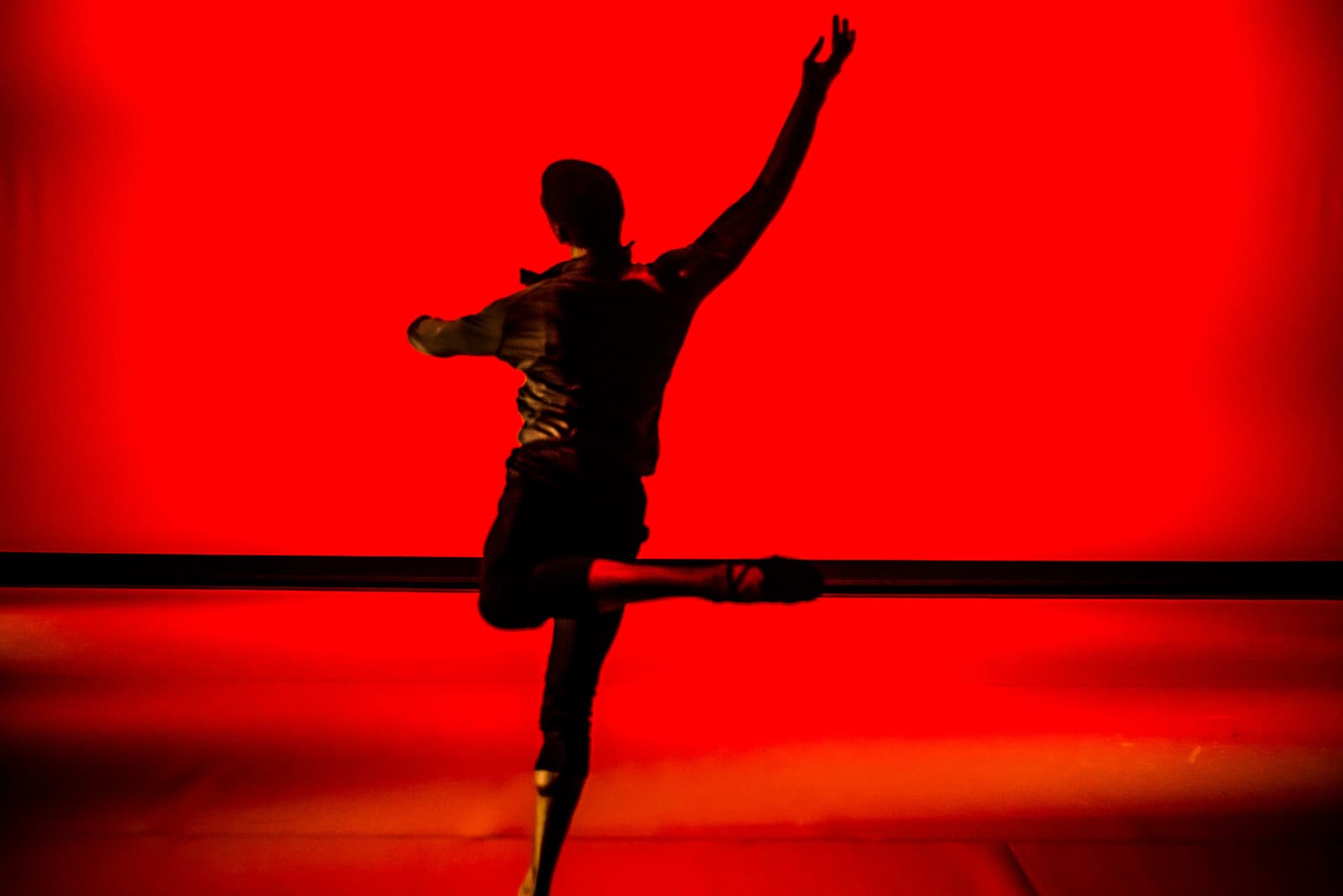 A dancer performing a ballet move against a vibrant red backdrop. The person is captured as a silhouette, arms raised and one leg extended behind them. A horizontal black line traverses the middle of the frame, marking a stage boundary.