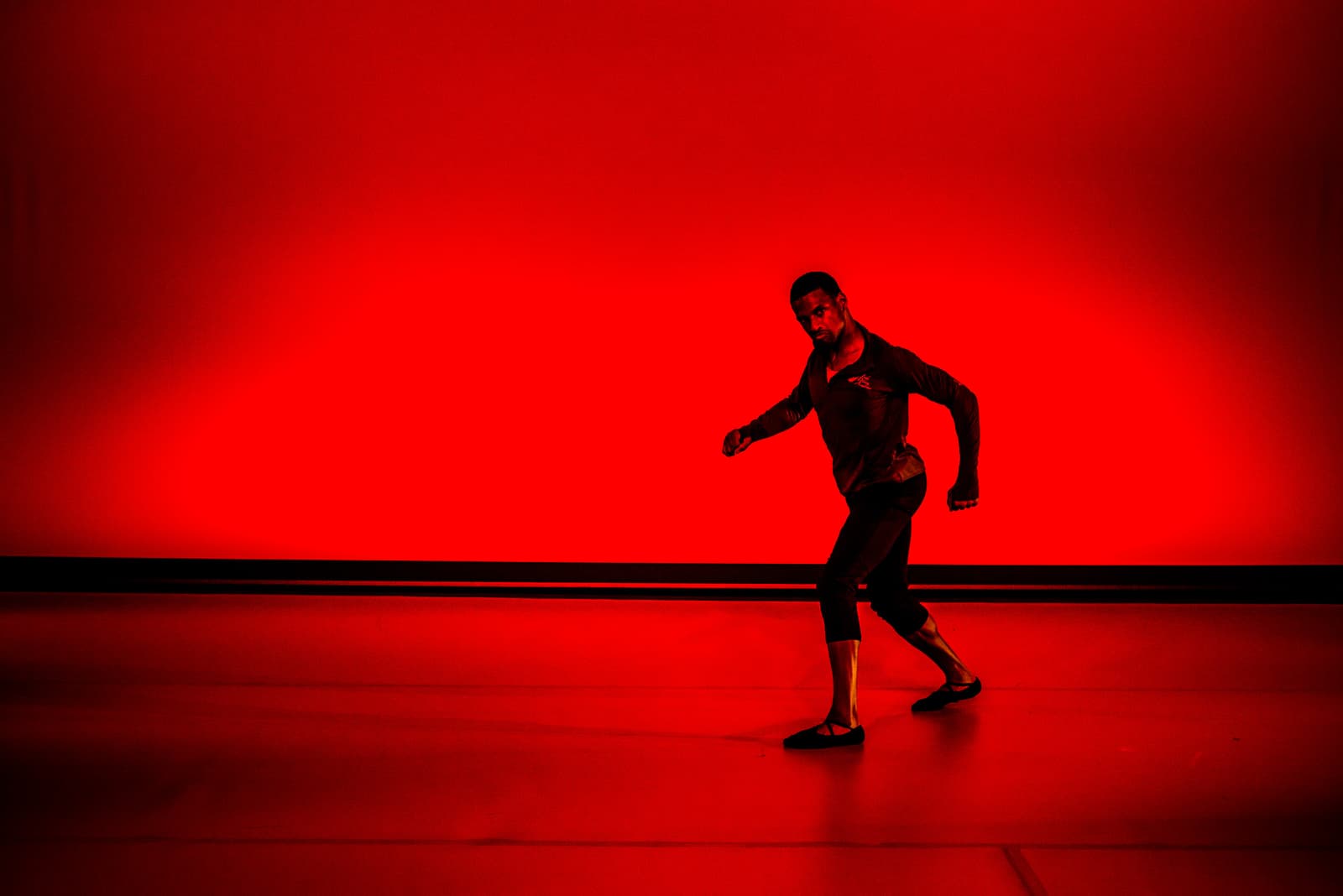 A dancer in dark form-fitting clothing performs on a stage lit by intense, saturated red light. The dancer is captured in a wide stance, turned slightly toward the viewer, with one arm extended outward and the other bent at the elbow. The background is a flat, solid, luminous red backdrop, and the stage floor reflects the bright red hue.