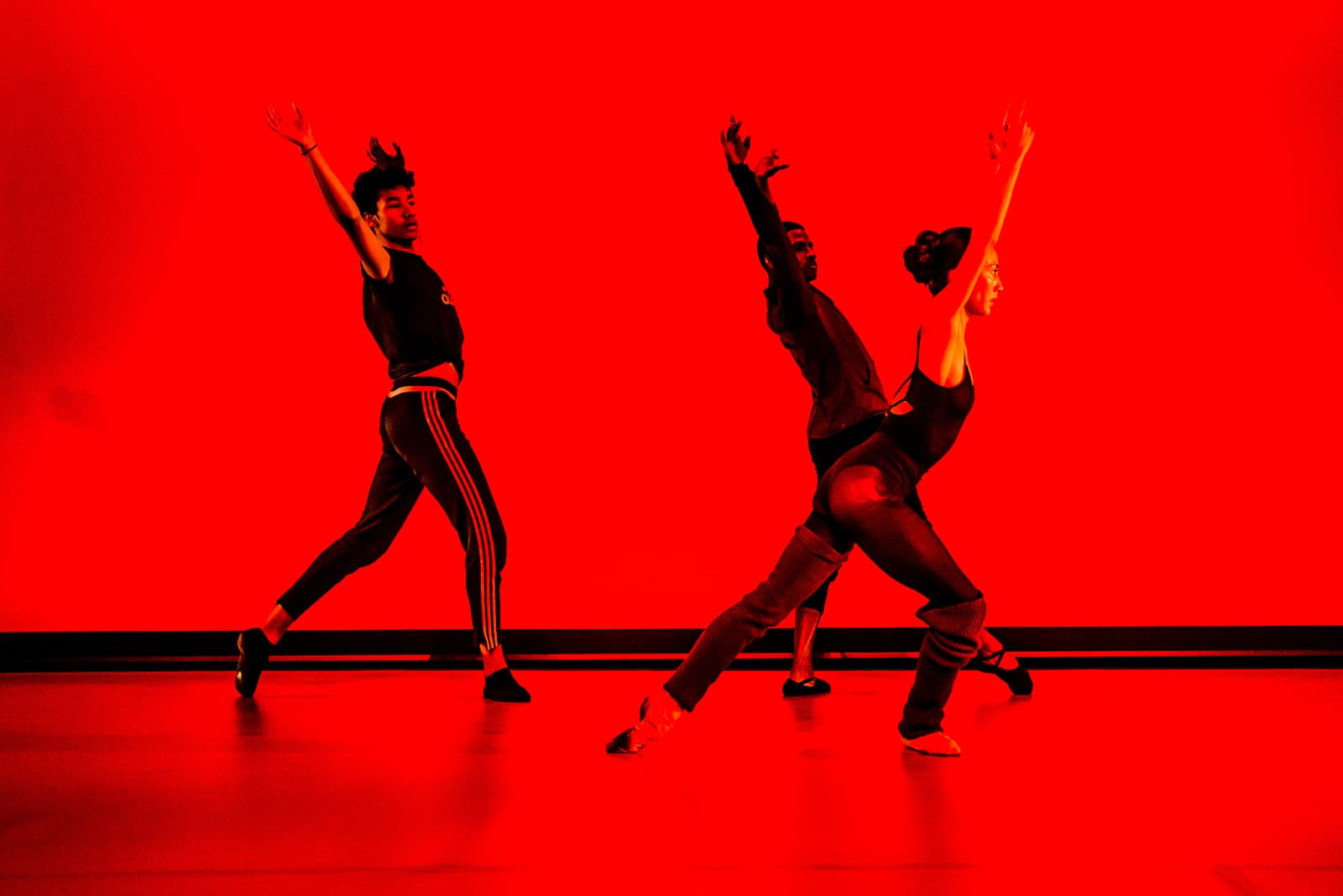 Three dancers perform on a stage illuminated by deep red light. One dancer stands on the left with one arm raised. To the right, two other dancers are in a synchronized pose, with one leaning forward while extending both arms upward. They wear dark exercise clothing and dance shoes against a solid, brightly lit red background.