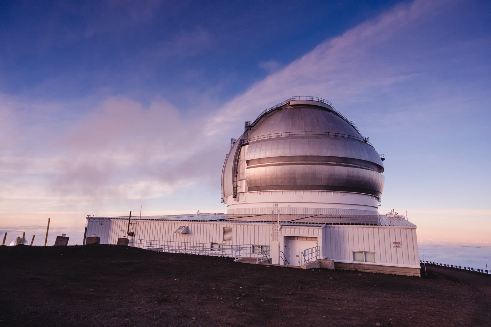 A large, metallic, domed astronomical observatory building sits atop a dark volcanic mountain summit above a thick layer of clouds. The sky shows soft pink and blue hues of dawn or dusk.