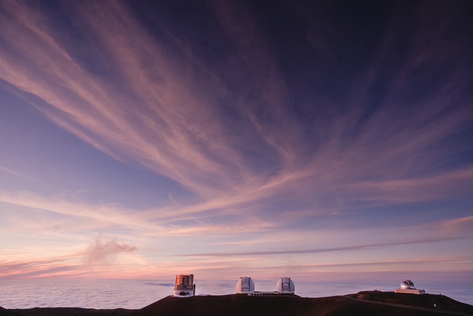 A wide angle view of several astronomical observatory domes situated on a dark volcanic ridge. Below the ridge, a thick layer of clouds covers the landscape, illuminated by a sunset with streaks of orange, pink, and purple in the sky.