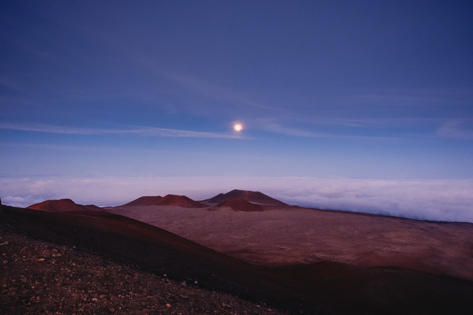 A wide landscape view of red volcanic craters under a twilight sky with a bright moon visible. A dense layer of clouds sits below the volcanic peaks, stretching to the horizon.