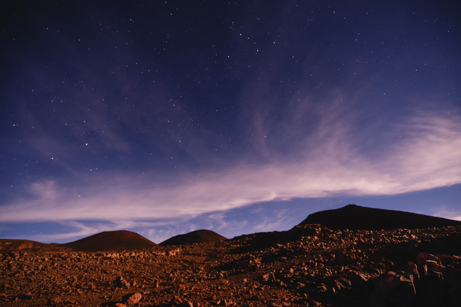 A wide angle night landscape of a rocky volcanic summit with reddish brown basalt stones in the foreground. Three dark, smooth volcanic mounds rise against a dark blue sky filled with many small stars and thin, wispy clouds illuminated by moonlight.