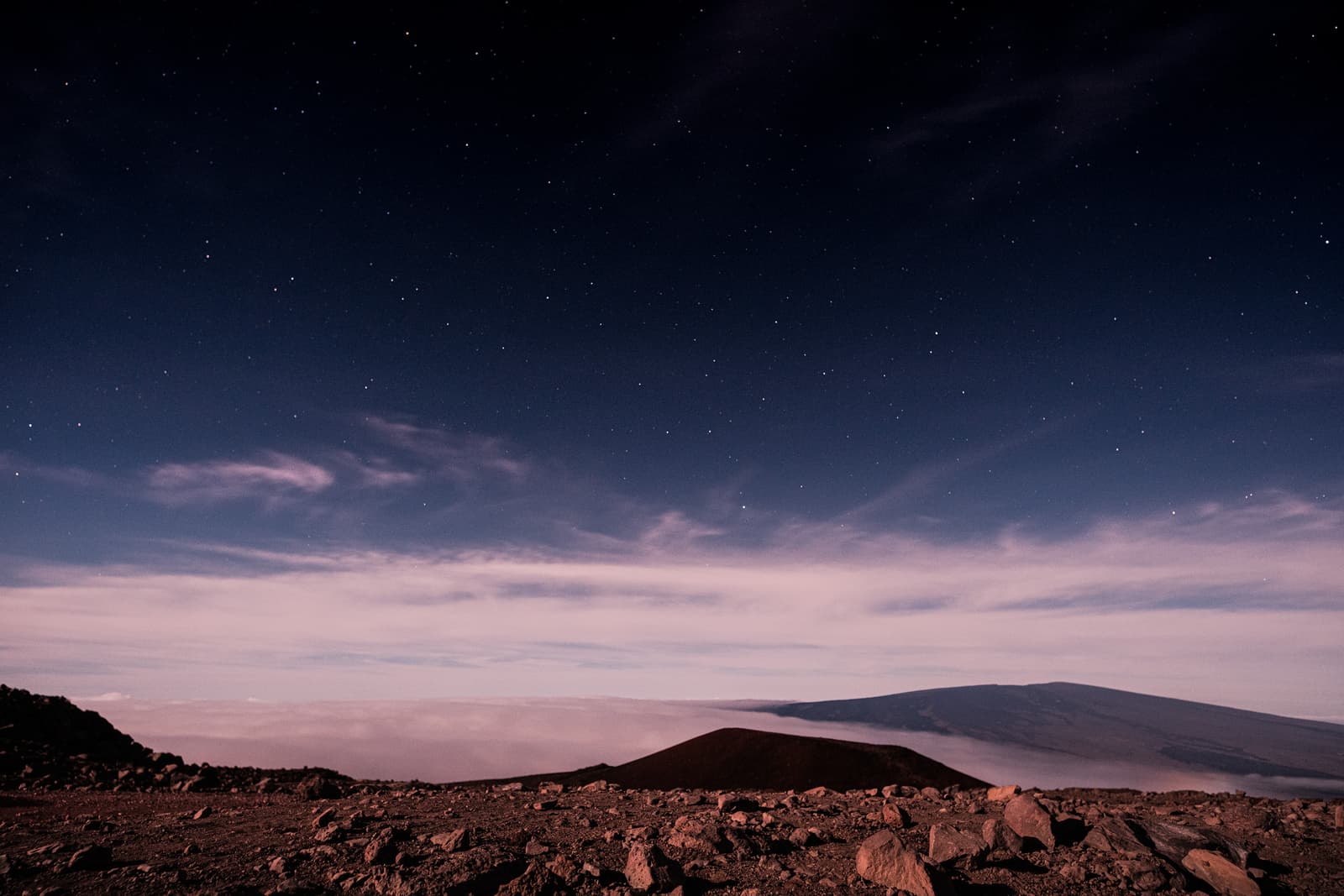 A wide-angle night landscape view from a rocky, dark volcanic mountain peak overlooking a thick layer of clouds below. In the distance, a large mountain silhouette rises above the cloud line. The sky is dark blue and filled with scattered stars. The foreground is composed of sharp, loose dark volcanic rocks illuminated by dim, ambient light.