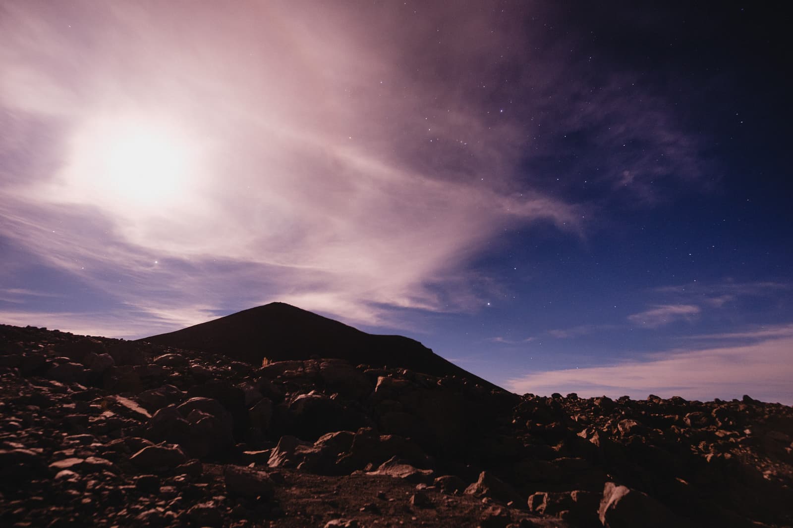 A wide angle night view of a volcanic peak under a starry sky. The foreground is composed of jagged, dark basalt rocks. The sky features wispy clouds illuminated by moonlight, casting a soft, purple-hued glow over the landscape.