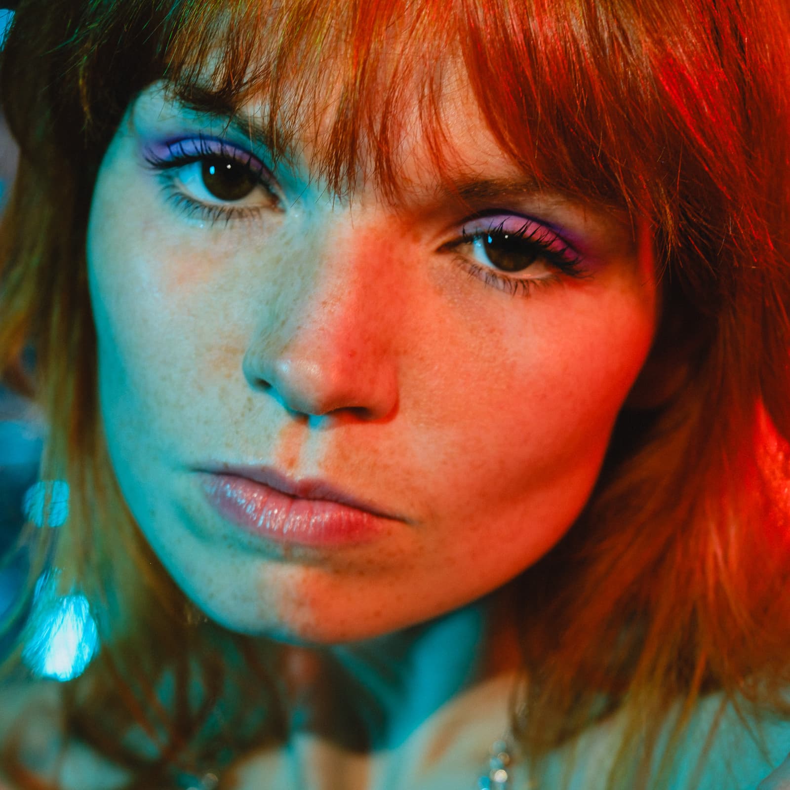 A tight, close-up portrait of a person looking directly into the camera. The face is illuminated by contrasting red and teal light, casting distinct colour shadows across their skin. They have copper-toned hair and are wearing vibrant purple eyeshadow.