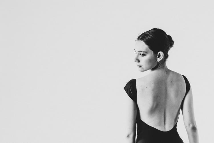 A black-and-white medium shot of a person facing away from the camera, looking over their right shoulder. They are wearing a plain black leotard with a deep scoop back and their hair is pulled back into a neat bun against a neutral light gray studio background.