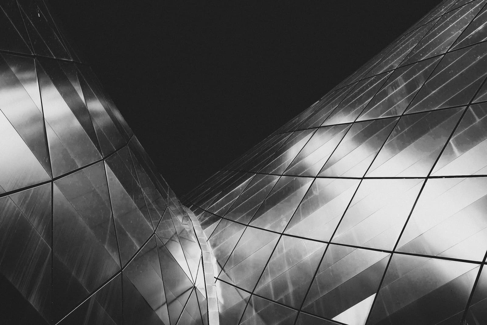 A black-and-white low-angle close-up of a modern building facade featuring a curved, faceted steel skin. Triangular metal panels meet at sharp joints, reflecting light in a gradient of bright whites to deep grays, with a solid black void in the upper center of the frame.