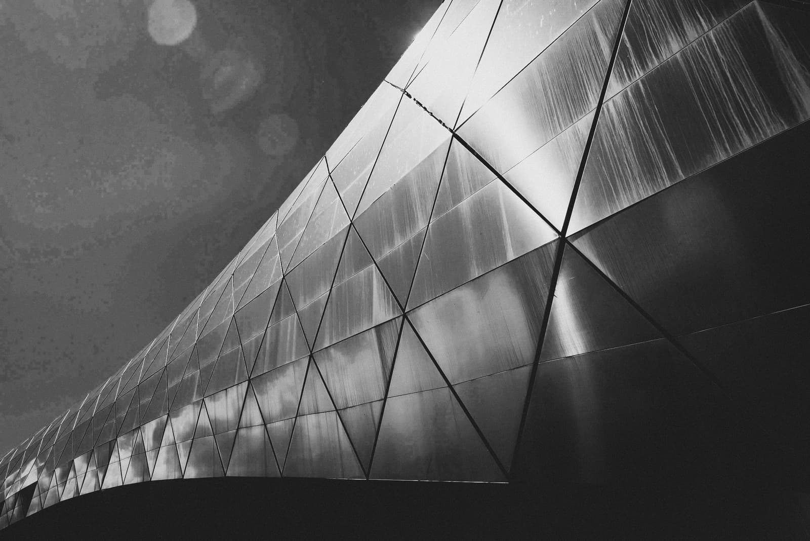 A black-and-white low-angle close-up of a modern building facade featuring a repeating diamond-shaped glass and metal lattice structure reflecting light. The surface shows vertical streaks and gradients of grey, transitioning from a brightly lit diagonal edge to deep shadows at the base.
