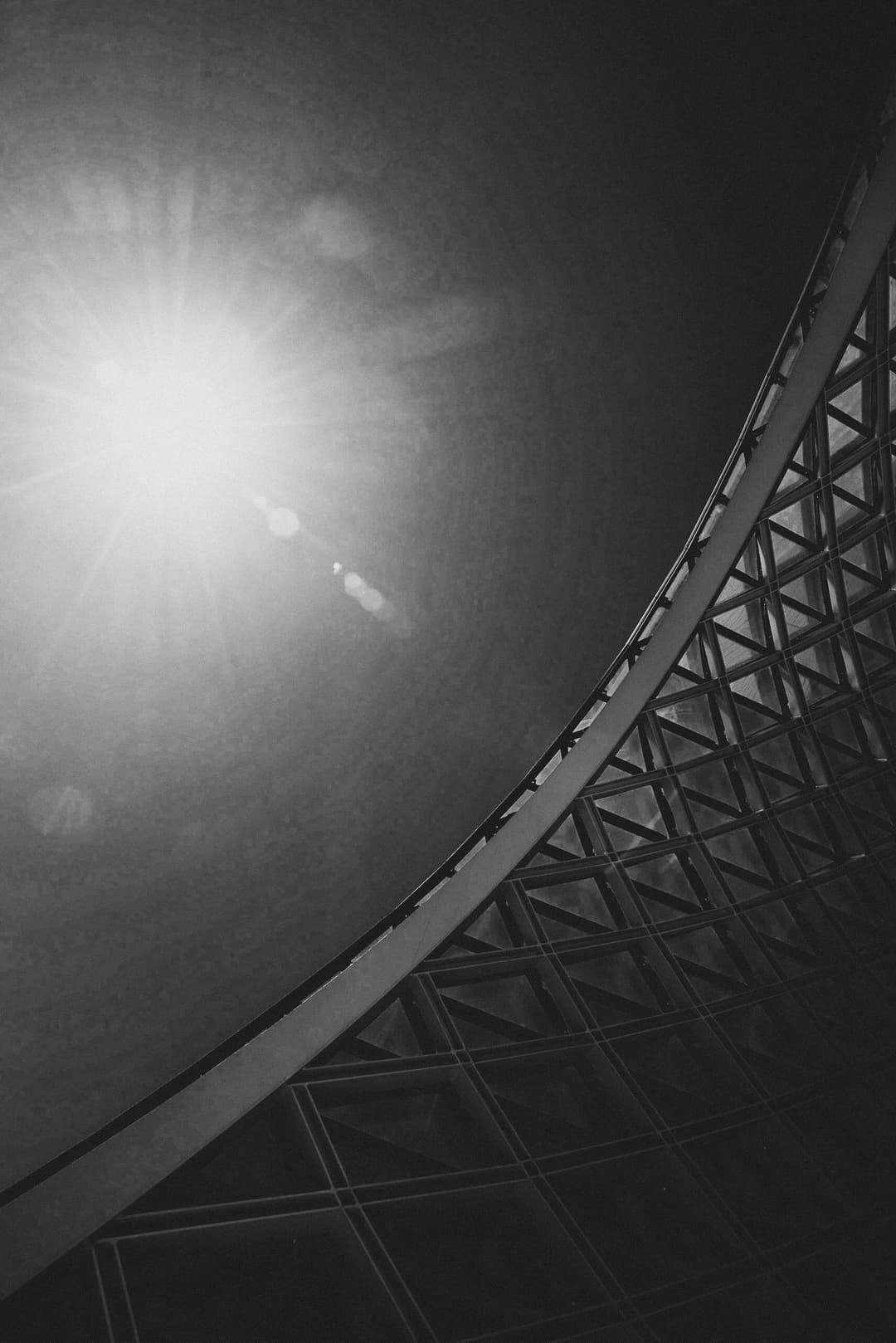 A black and white low-angle shot of a curved architectural structure with a metal triangular grid pattern. A bright sun flares in the upper left corner, creating a lens flare against the dark sky. The curved edge of the building cuts diagonally across the frame.