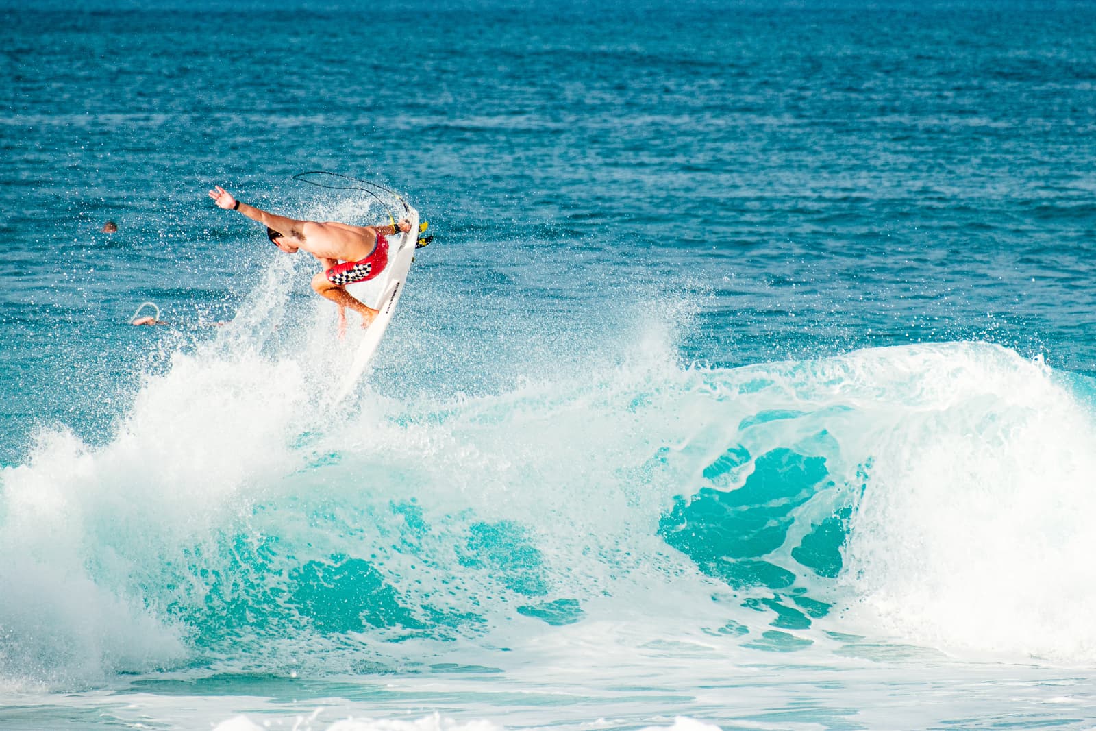 A surfer performs an aerial maneuver above the crest of a breaking wave at Pipeline, with the turquoise water exploding into sea spray around the white surfboard.