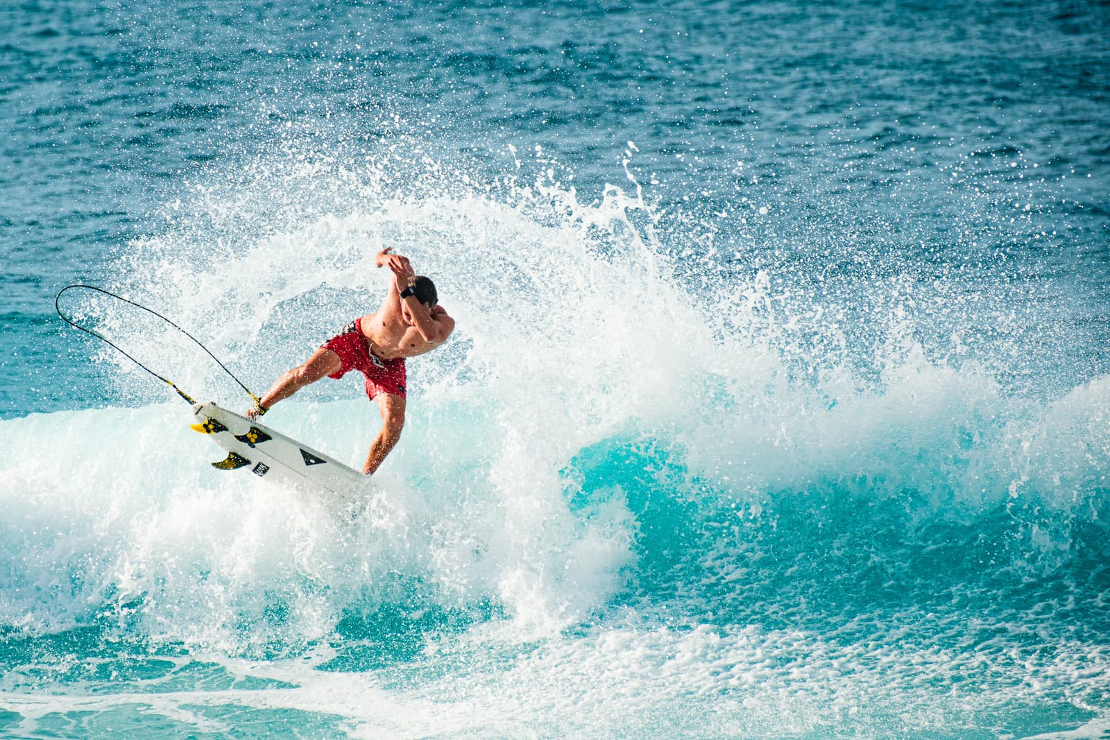 A shirtless person wearing red swim shorts rides a white surfboard across a bright turquoise ocean wave. The person is mid-turn with their body angled toward the water, creating a large, dramatic splash of white sea foam behind them. The surfboard has black fins and a thin black leash trailing through the air.