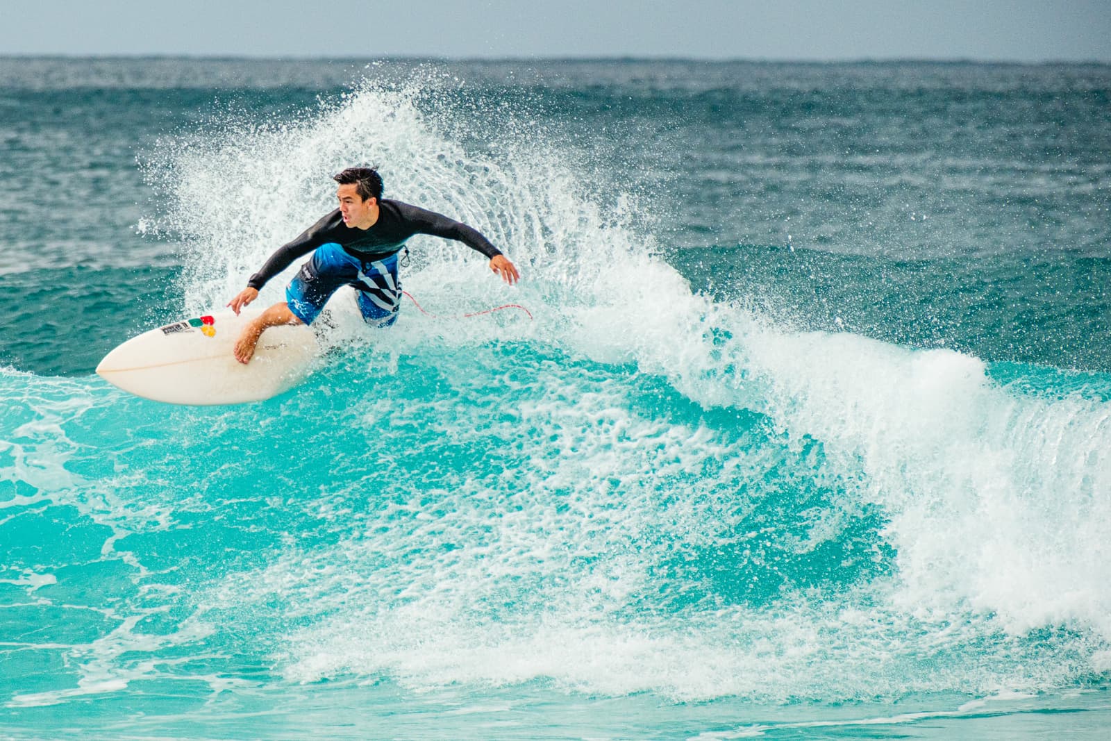 A surfer in a black long-sleeve rash guard and patterned blue board shorts rides a turquoise wave at Banzai Pipeline, North Shore, Oahu. The surfer is crouched on a white surfboard, kicking up a large spray of water behind them, with the ocean extending to the horizon.