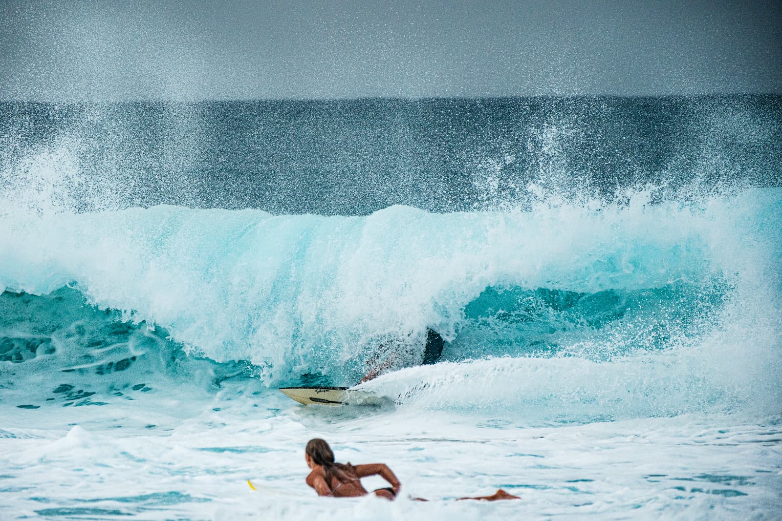 A surfer rides a large, curling wave at Banzai Pipeline, North Shore, Oahu. The crest of the wave is bright turquoise and translucent, with white sea spray catching the light. In the foreground, a person with long hair lies on a surfboard, facing toward the breaking wave.
