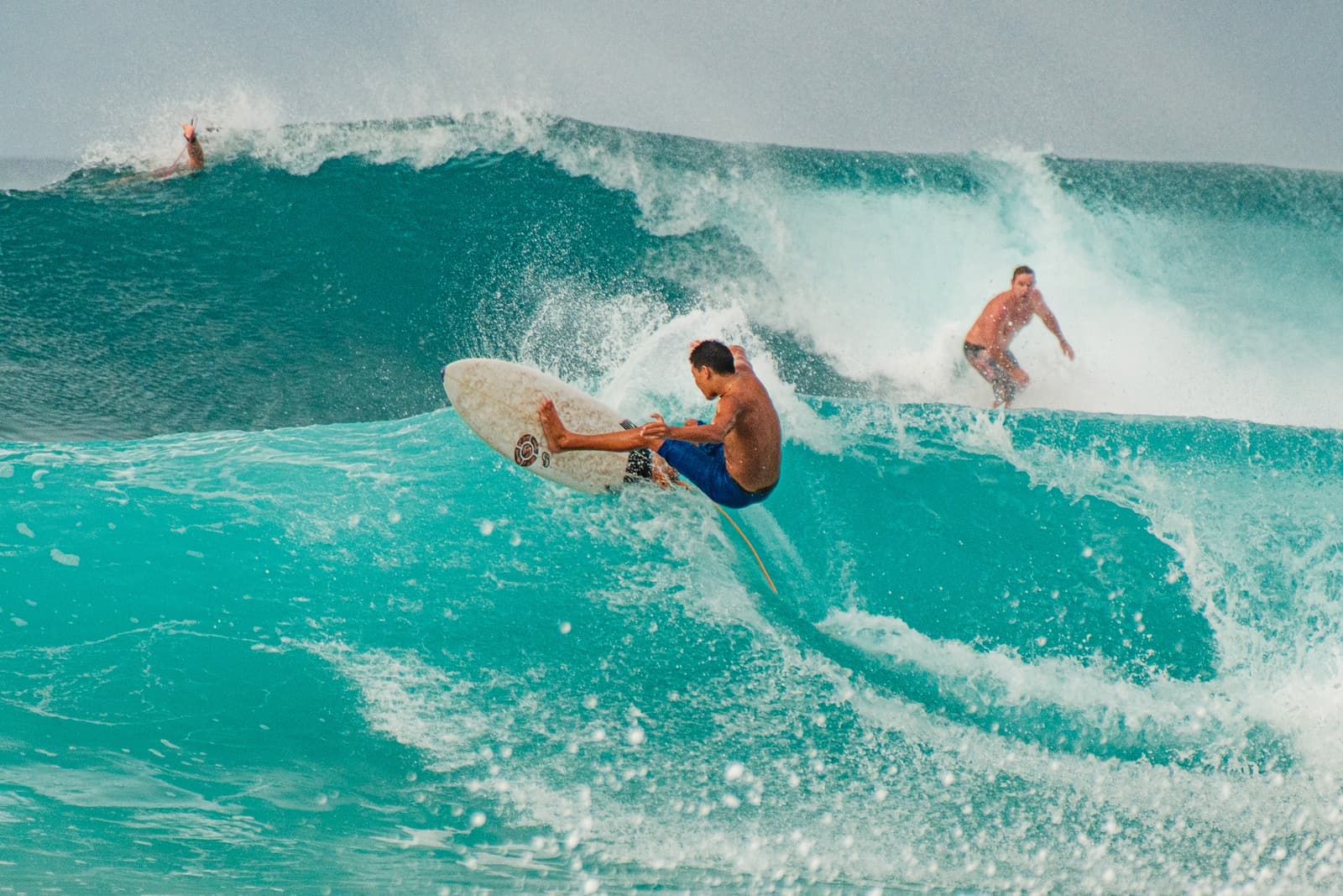 A surfer rides a bright turquoise wave while performing a sharp turn on a surfboard. Another surfer is visible in the background, riding a separate part of the same wave, while another person's limbs are visible emerging from the crest of the breaking wave behind them, all set against an overcast sky.