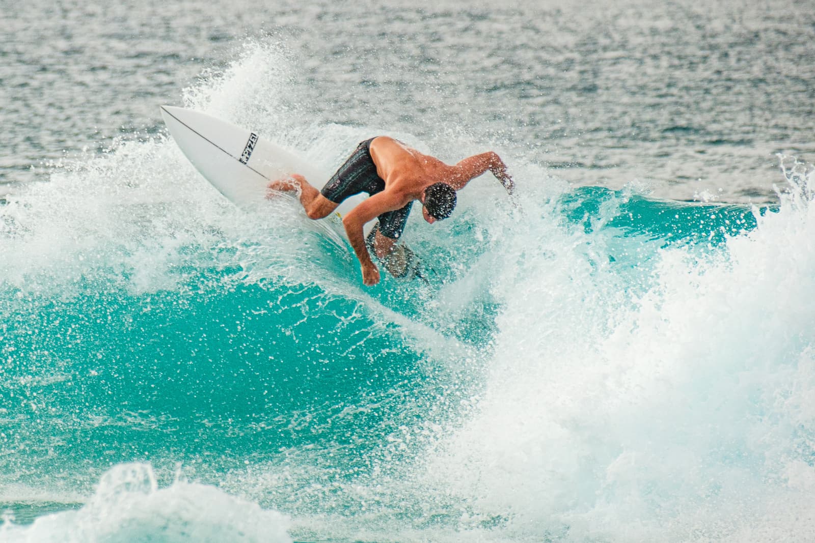 A surfer in a low crouch leans into a sharp turn on a turquoise wave, white sea spray exploding around the board. The surfer is bare-chested and wearing dark boardshorts.