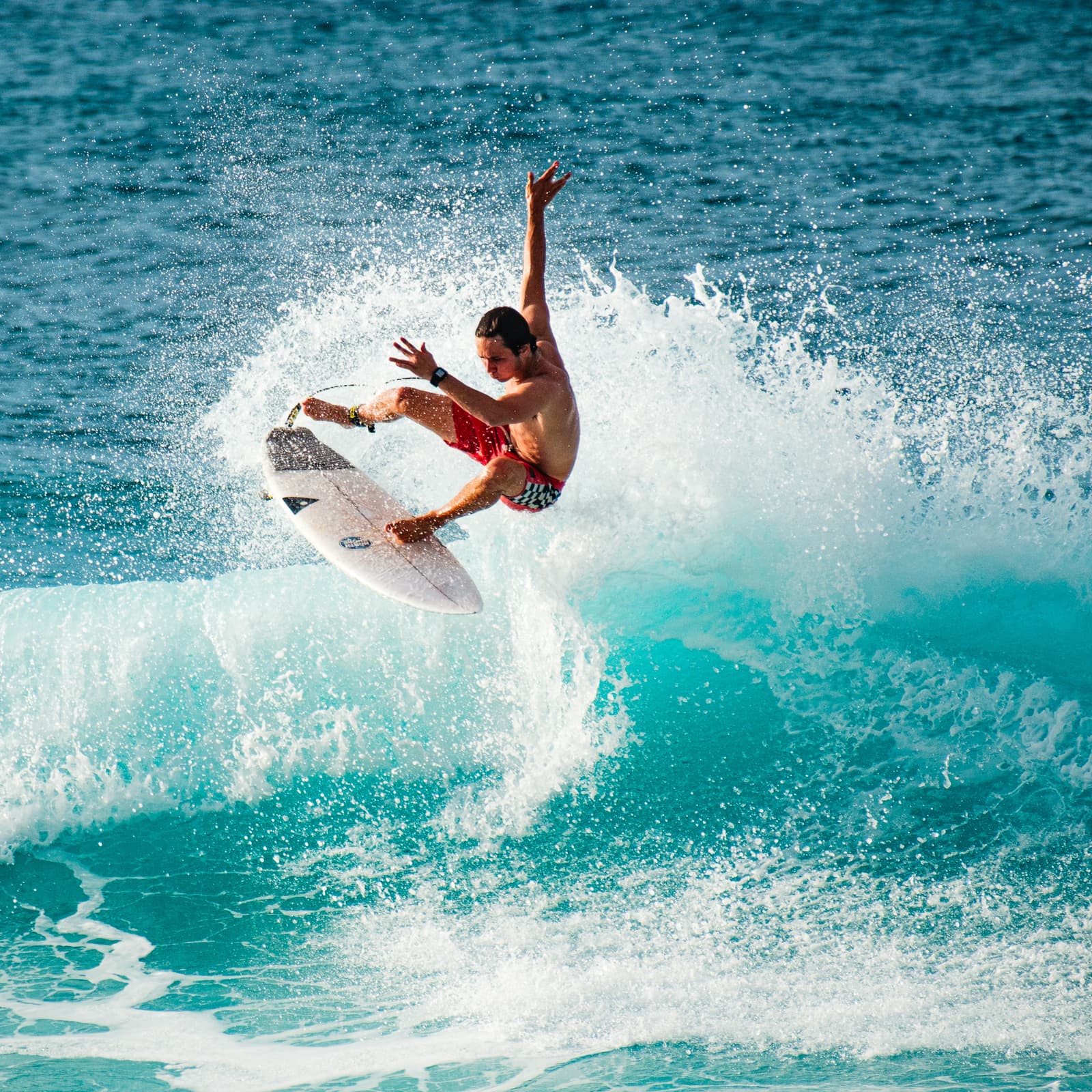 A person airborne on a surfboard above a breaking wave at Banzai Pipeline. The subject is mid-air, body angled with one arm extended upward and the other reaching toward the board. They are wearing red and white patterned swim trunks. Ocean spray and white water surround the board and the surfer against a bright blue sea.