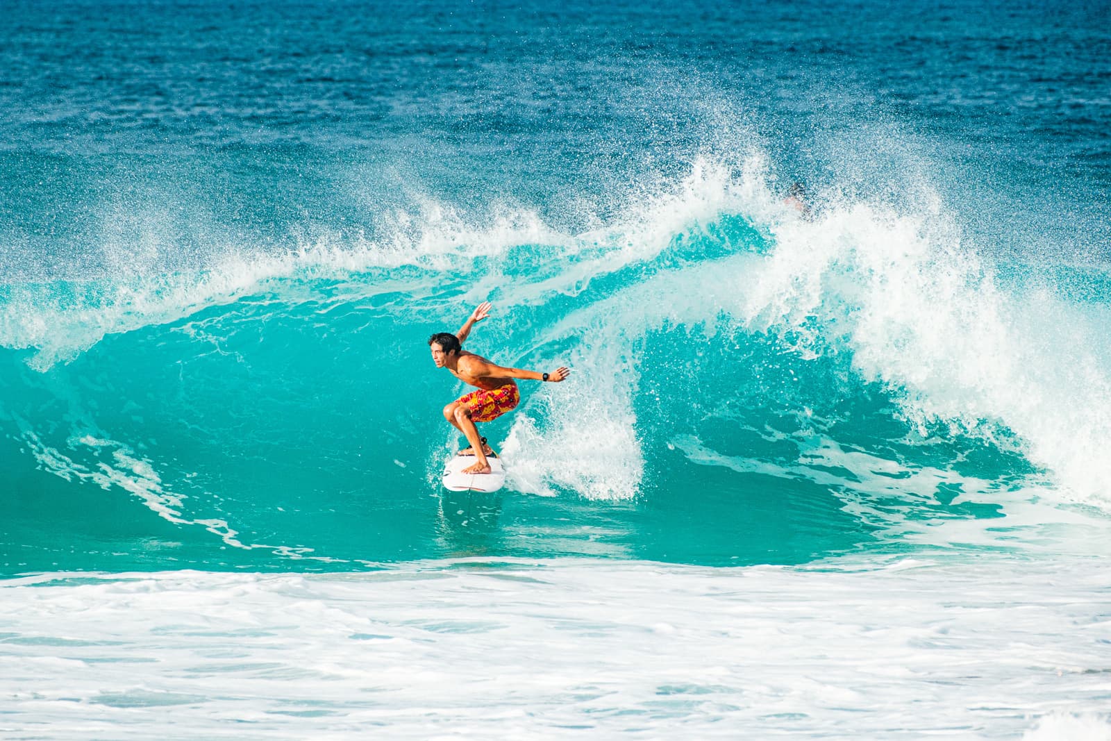 A person surfing on a large, turquoise wave at Banzai Pipeline. The surfer is wearing patterned orange boardshorts and is balanced on a white surfboard, one arm extended outwards. The water is bright blue with white sea spray curling over the top of the wave.