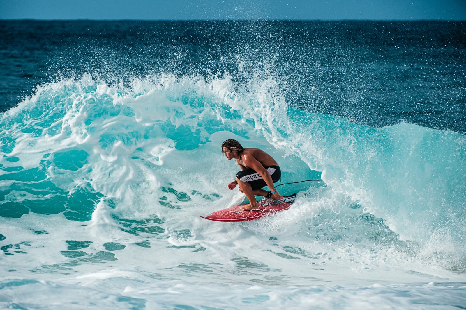 A surfer with long blonde hair crouched low on a red surfboard inside the tube of a large ocean wave, surrounded by turquoise water and white sea spray.