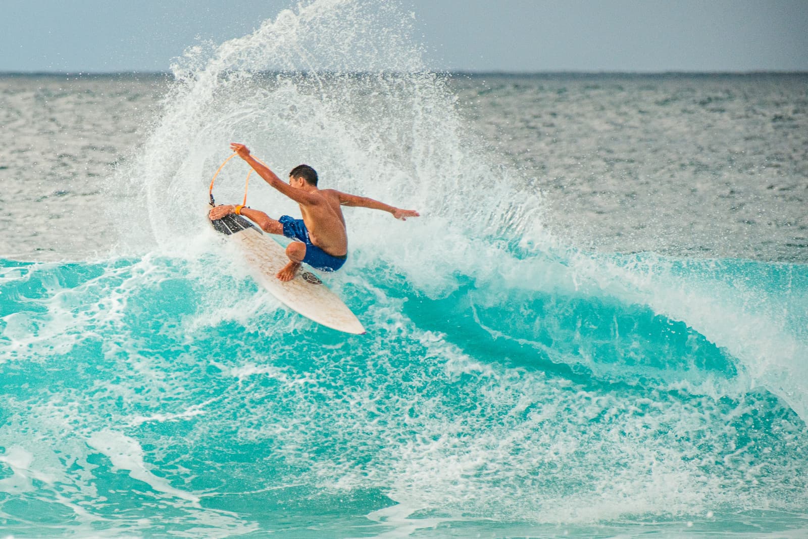 A person surfing on a cresting turquoise wave at the Banzai Pipeline, extending their arms and one leg outward to maintain balance as water sprays around them, set against a horizon line of the open ocean.