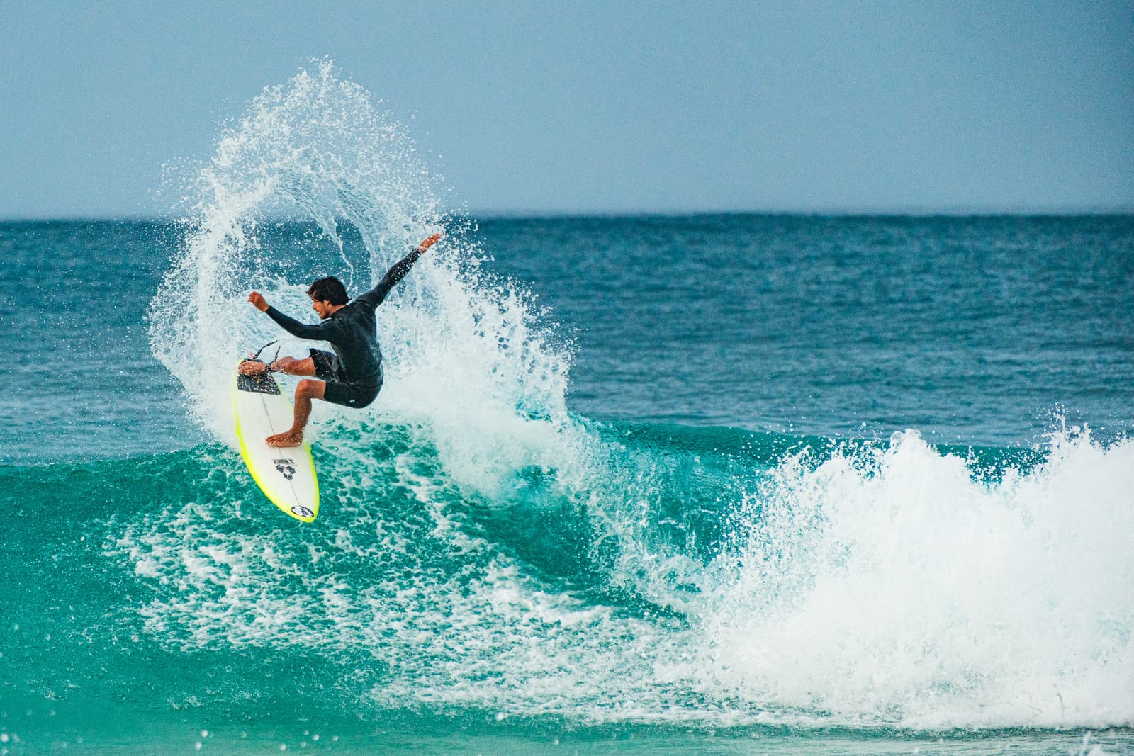 A surfer in a dark wetsuit performing a high-speed turn on a turquoise wave, carving upward as a large spray of water arcs behind them like a halo. The surfboard is yellow and white, and the background is an open, deep blue ocean under bright daylight.