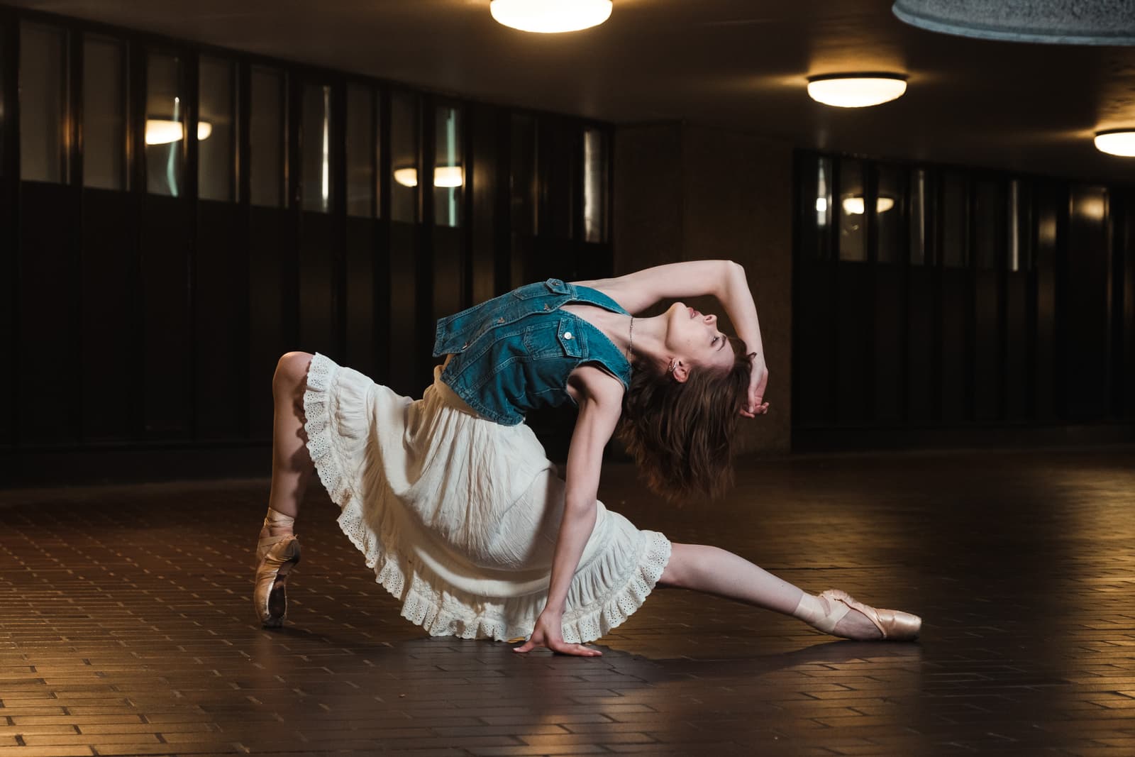 A person in a blue denim vest and a white tiered skirt is captured in a deep, arched dance pose on a dark tiled floor. The person's left leg is bent behind them with a ballet slipper touching the floor, while the right leg is extended forward. Their back is arched, head tilted back, with one arm overhead and the other resting on the floor for support. The background is a dark, enclosed space with rows of dim, recessed ceiling lights reflected in polished dark walls.