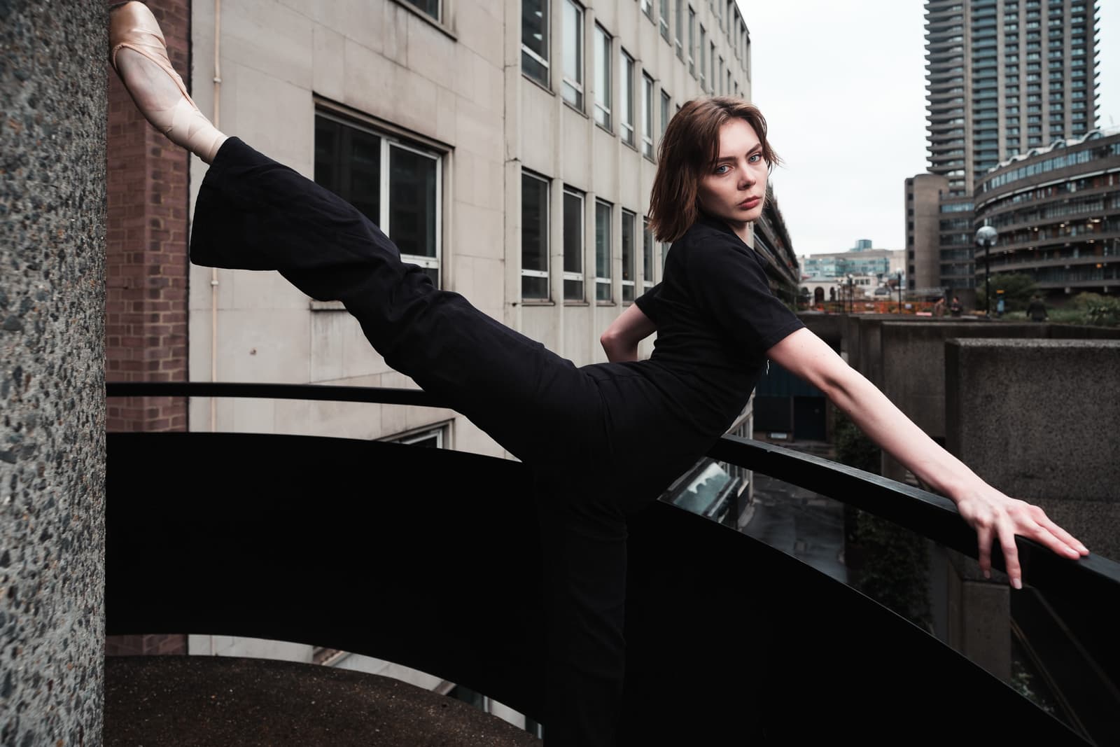 A person with short hair wearing a black top and black trousers performs a high leg extension, with one pointe shoe resting against a concrete pillar and one hand resting on a black railing. The background is an urban concrete landscape of brutalist-style buildings on an overcast day.