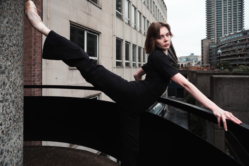 A person with short hair wearing a black top and black trousers performs a high leg extension, with one pointe shoe resting against a concrete pillar and one hand resting on a black railing. The background is an urban concrete landscape of brutalist-style buildings on an overcast day.