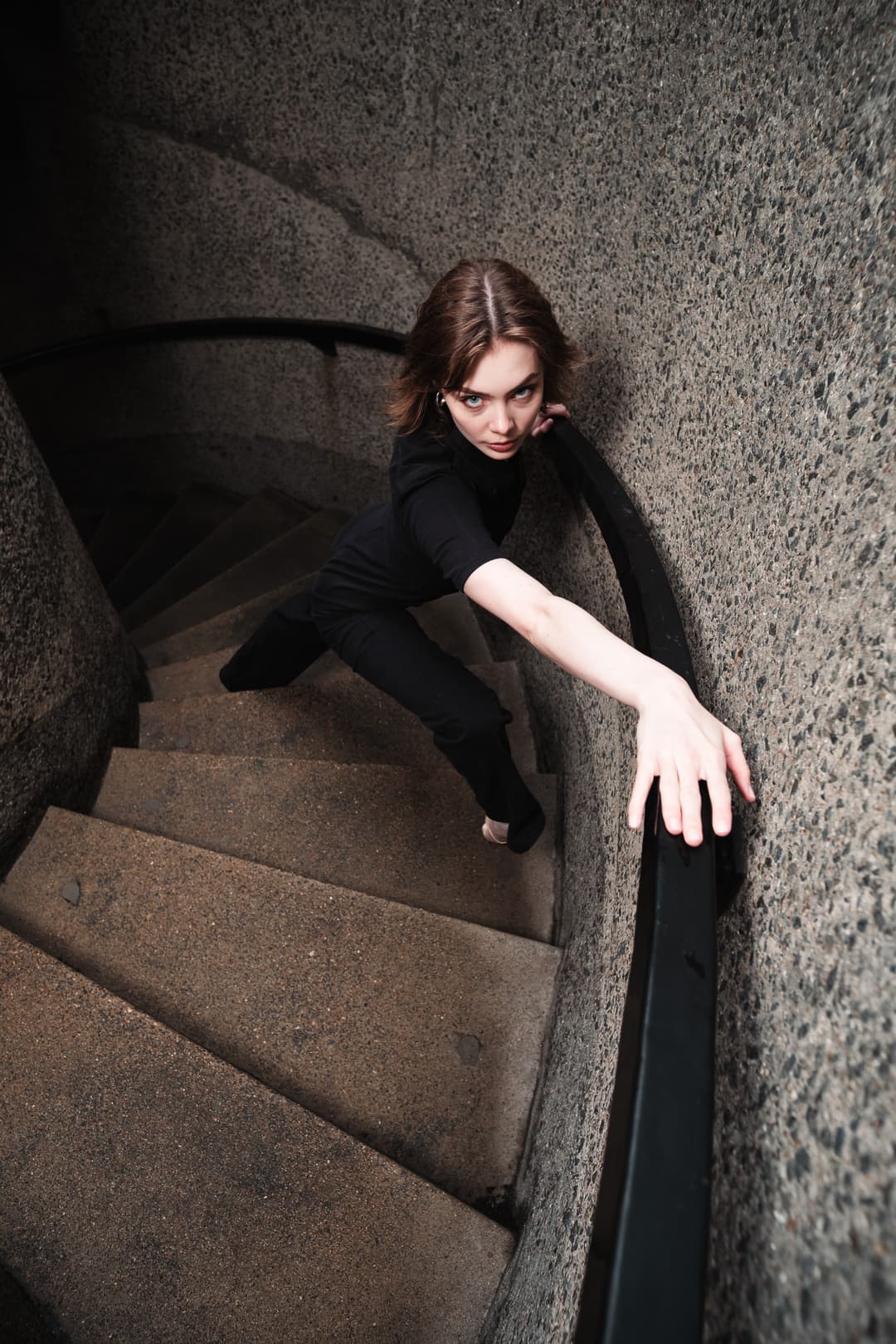 A high-angle view of a person wearing a black short-sleeved top and dark trousers, posed on a concrete spiral staircase. They are leaning forward with one arm extended along a curved black handrail, looking up directly toward the camera. The background is composed of rough, grey textured concrete walls and stairs.