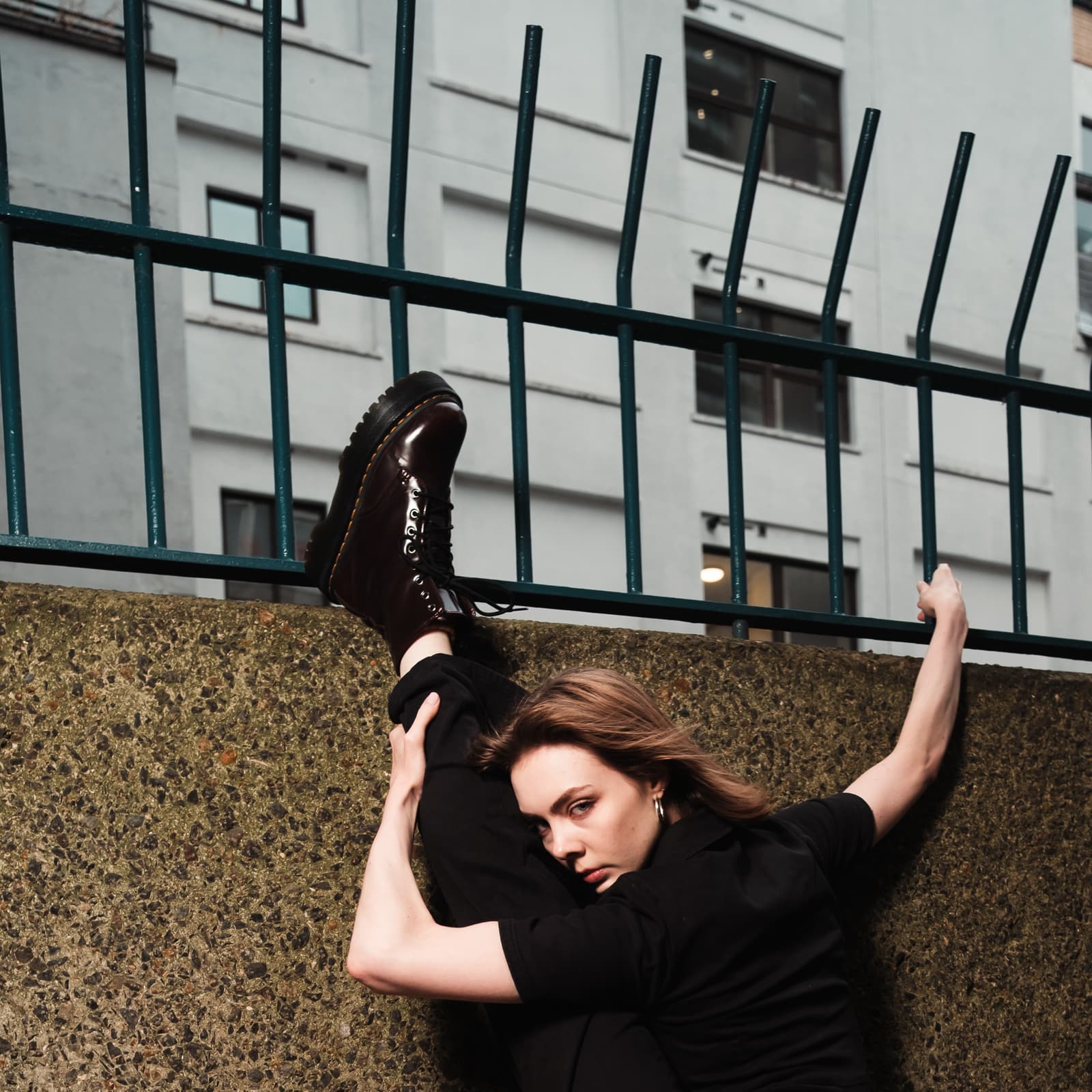 A person in black clothing holds a leg high in a vertical split against a textured concrete wall topped with a green metal fence. The person wears dark-colored, shiny lace-up boots and looks towards the camera, leaning against the fence for balance in an outdoor urban setting.