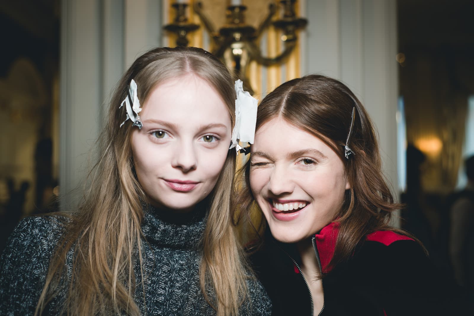 A close-up photograph of two individuals in an indoor setting. The person on the left has long, light brown hair with white paper hair-setting strips clipped to the sides, wearing a grey knit turtleneck sweater. The person on the right has long dark hair with a silver clip, wearing a black and red zip-up jacket, and is winking with their tongue slightly visible. The background shows a blurry interior wall with a gilded, ornate wall sconce.