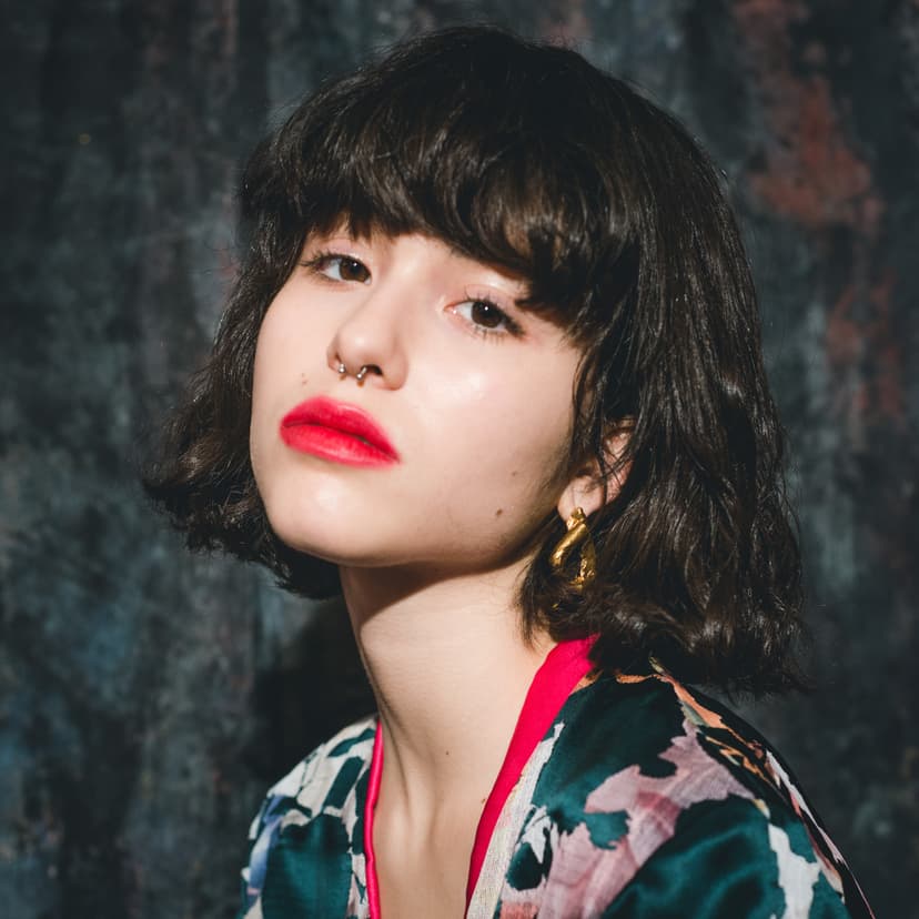 A close-up portrait of a person with chin-length dark hair and bangs, wearing a vibrant, floral-patterned silk garment with pink piping. They have a septum piercing and gold sculptural earrings, and their lips are painted a bold, matte red. They are positioned against a dark, mottled grey wall, illuminated by direct, high-contrast light.