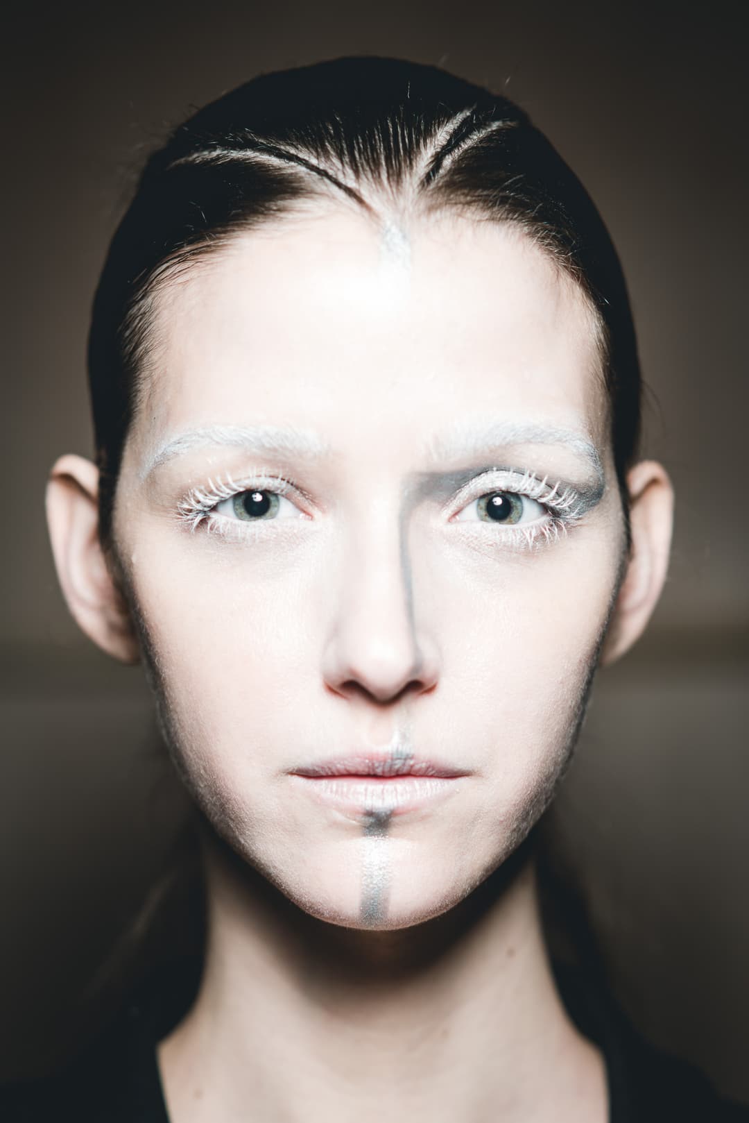 A close-up, frontal portrait of a face with very pale, almost white skin tone and dramatic makeup. The eyebrows and eyelashes are coated in white. A sharp, vertical grey line is painted down the center of the forehead, nose, and chin. The hair is pulled tightly back in a dark, sleek style. The background is a neutral, slightly out-of-focus grey-brown.