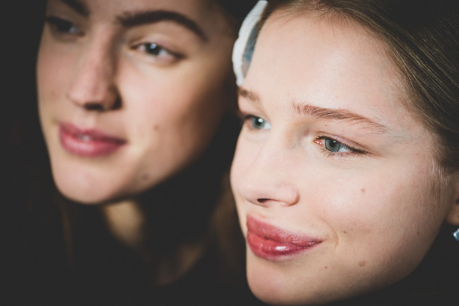 Close-up portrait of two individuals. One is in focus in the foreground on the right, looking slightly away from the camera with a subtle smile. A small white hair clip is visible on the side of their head. The second person is blurred in the background to the left, also looking in the same direction. The lighting is soft and natural against a dark background.