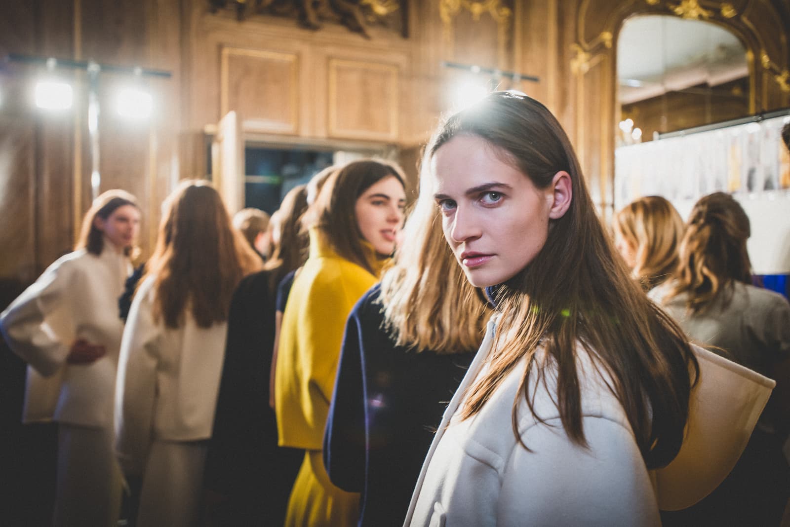 A close-up shot of a person with long brown hair glancing over their shoulder directly at the camera. They are wearing a structured cream coat. In the background, other individuals are visible in a dimly lit, ornately decorated room with bright stage lights visible in the distance, suggesting a backstage area at a fashion show.