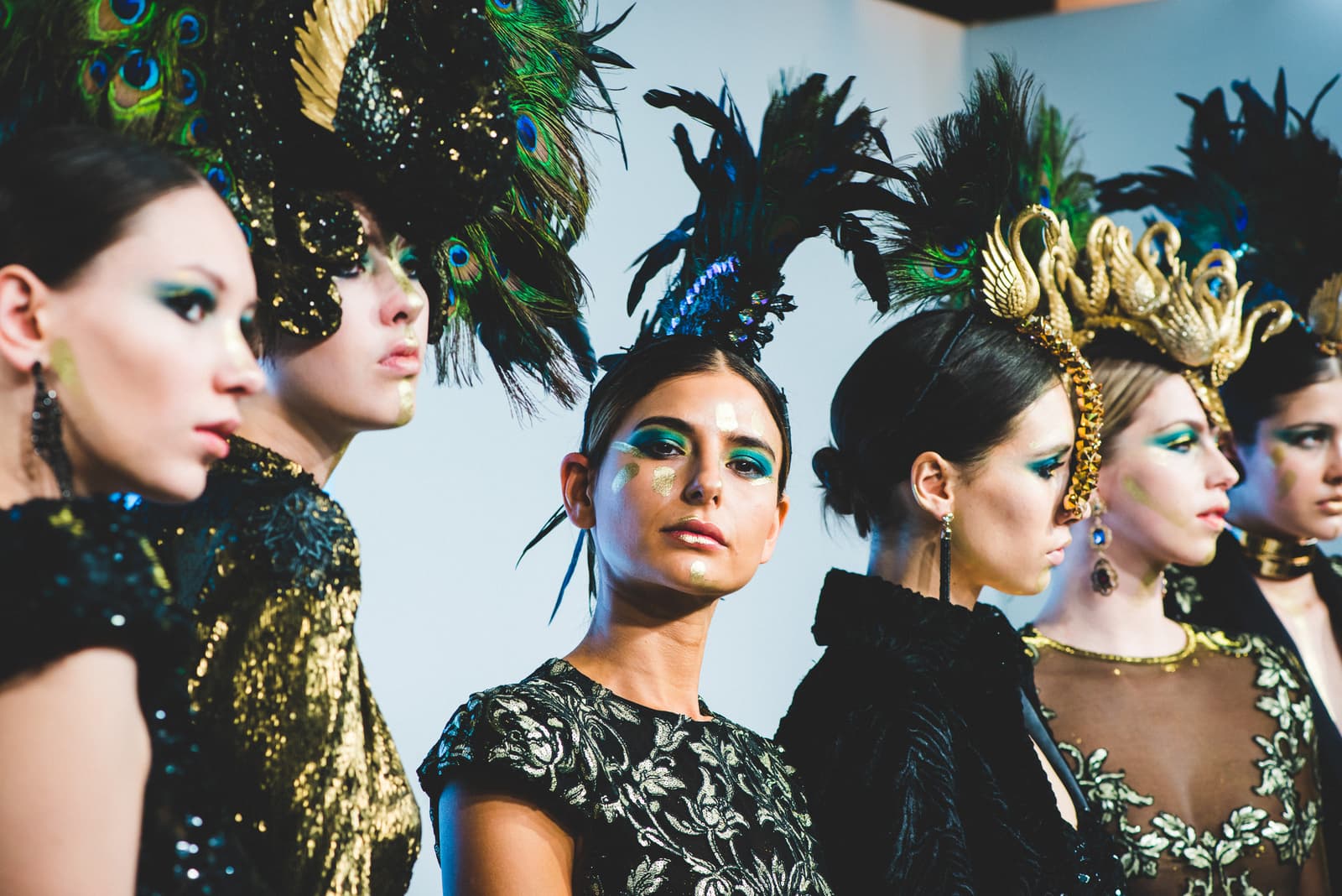 A line of models backstage wearing intricate headpieces featuring peacock feathers and gold structural elements, with matching emerald and gold makeup, dressed in elaborate textured and embroidered black clothing.