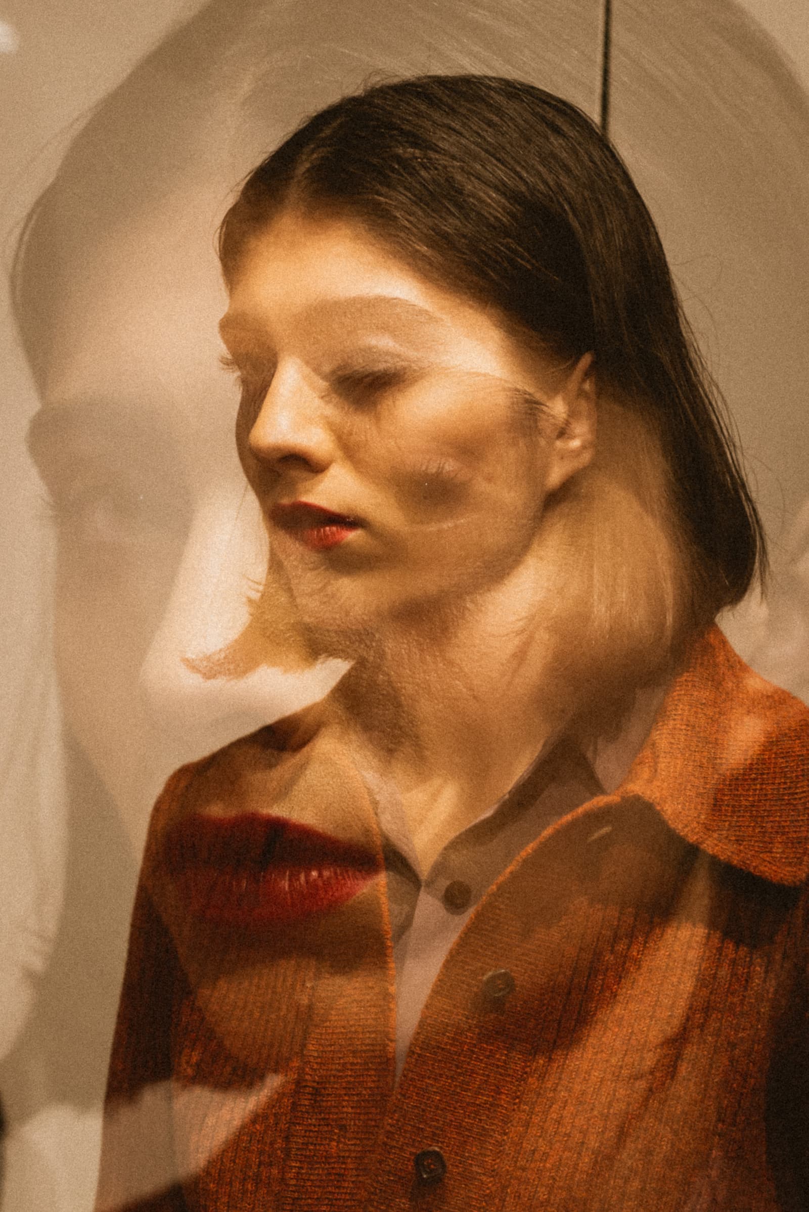 A double-exposed color portrait of a person in profile, facing left with eyes closed. A semi-transparent overlay shows a larger, ghostly version of the face in the background. The person wears a reddish-brown textured button-down shirt. The lighting is warm and sepia-toned, with a distinct red shape near the chin area.