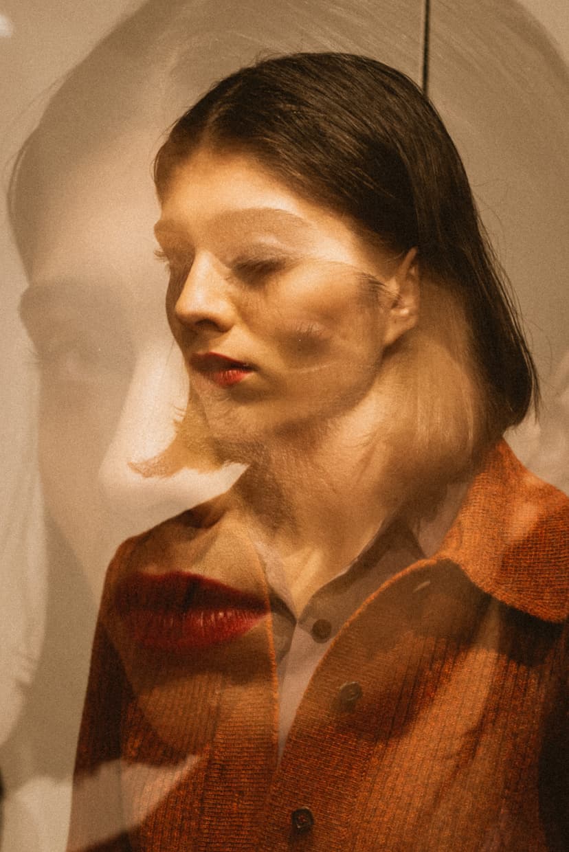 A double-exposed color portrait of a person in profile, facing left with eyes closed. A semi-transparent overlay shows a larger, ghostly version of the face in the background. The person wears a reddish-brown textured button-down shirt. The lighting is warm and sepia-toned, with a distinct red shape near the chin area.