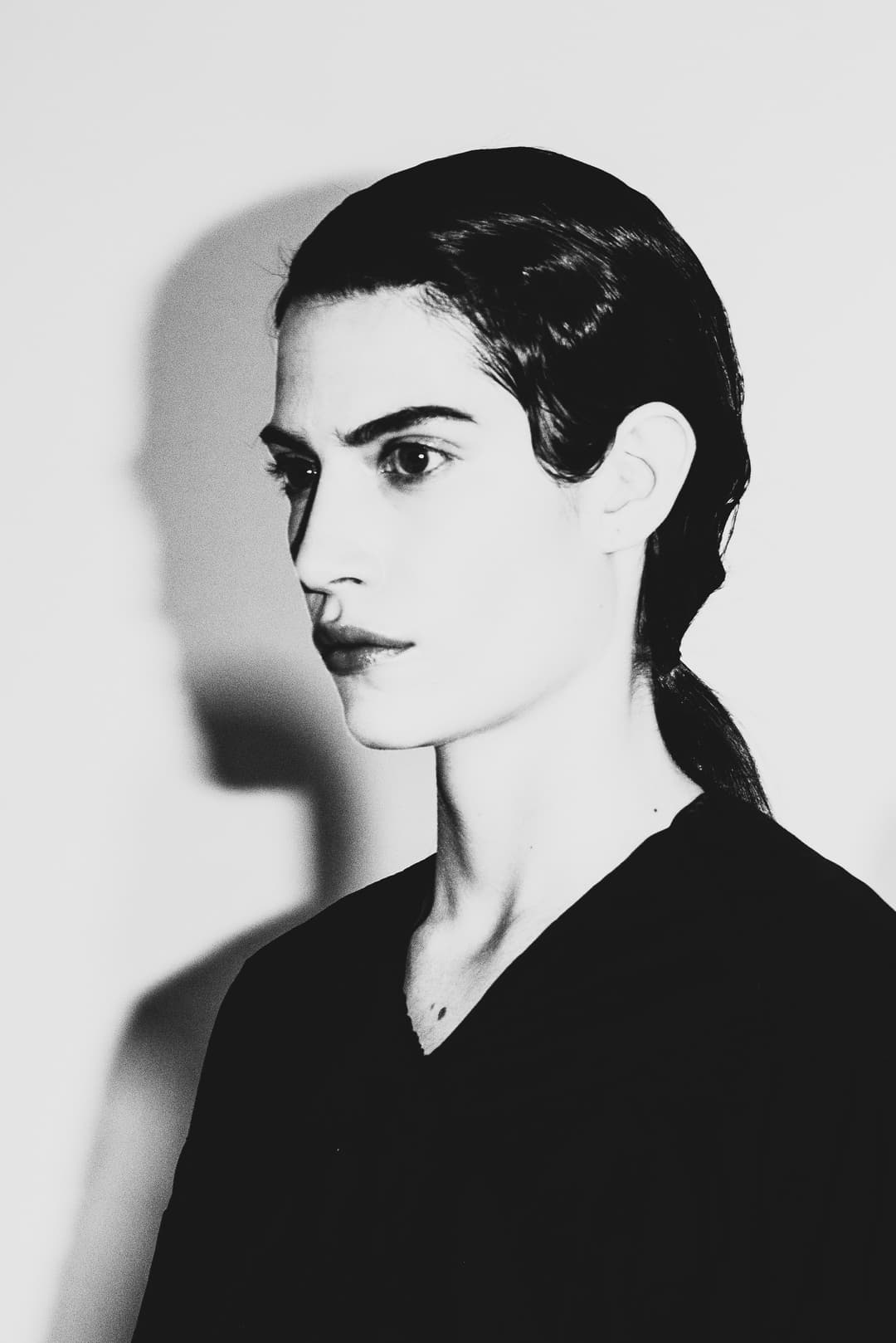 A black-and-white profile shot of a person with slicked-back dark hair, looking off-camera. The person wears a plain dark V-neck top. A sharp, dark shadow of their head and shoulders is cast against the plain white wall behind them, created by a strong light source from the side.