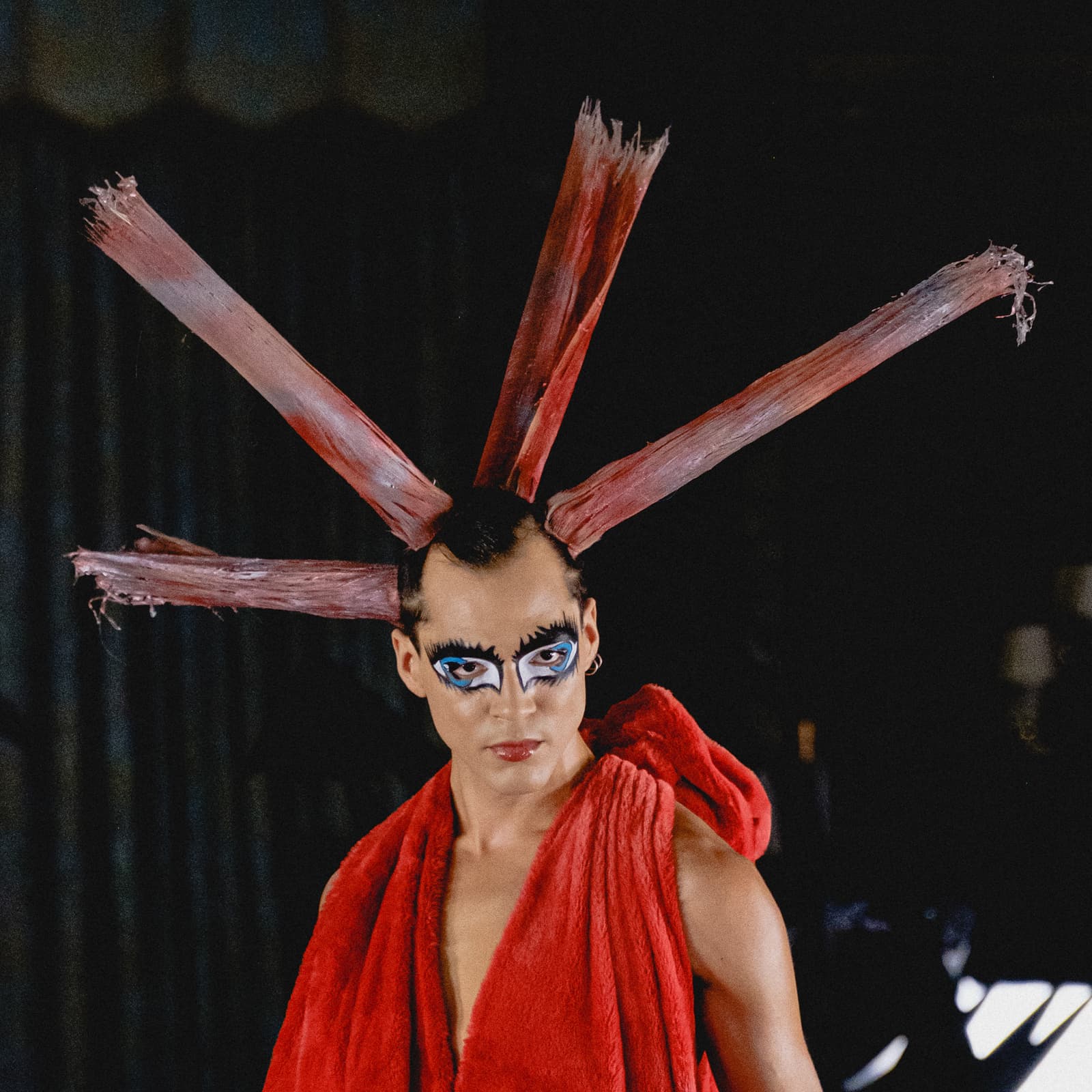 A person viewed from the chest up, wearing a bright red sleeveless garment with a textured, faux-fur appearance draped over the shoulders. The hair is styled into several stiff, radiating spikes of reddish-brown, mimicking the texture of dried bark or wood. The makeup features dramatic, dark graphic eyeliner and bright blue eyeshadow extending towards the temples. The background is dark and indistinct, suggesting a low-light indoor setting.