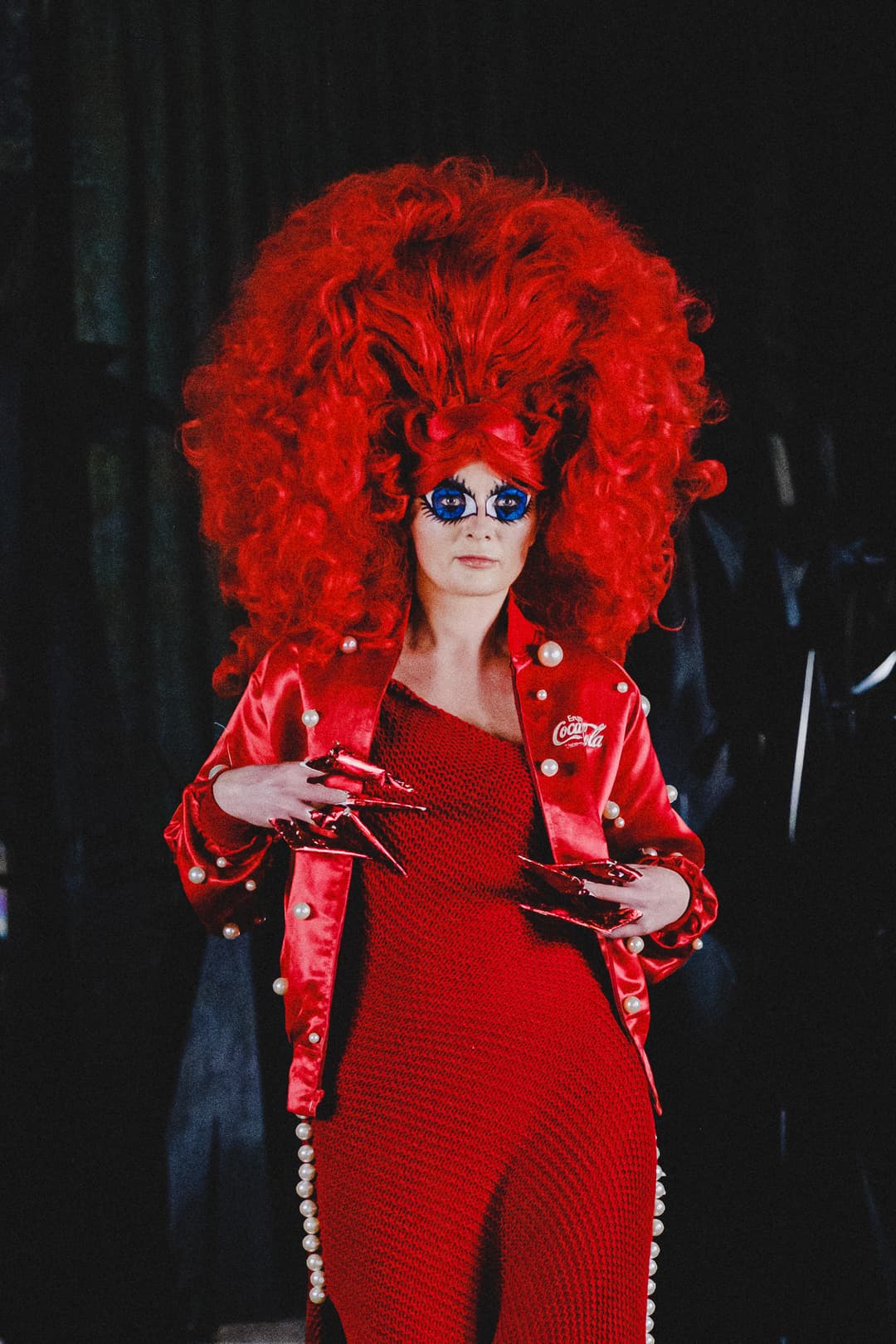 A person with voluminous bright red hair and large painted doll-like eyes gazes forward, wearing a red satin bomber jacket embellished with white pearls and a red knitted dress. The person has long, sharp red fingernails, set against a dark, out-of-focus background.