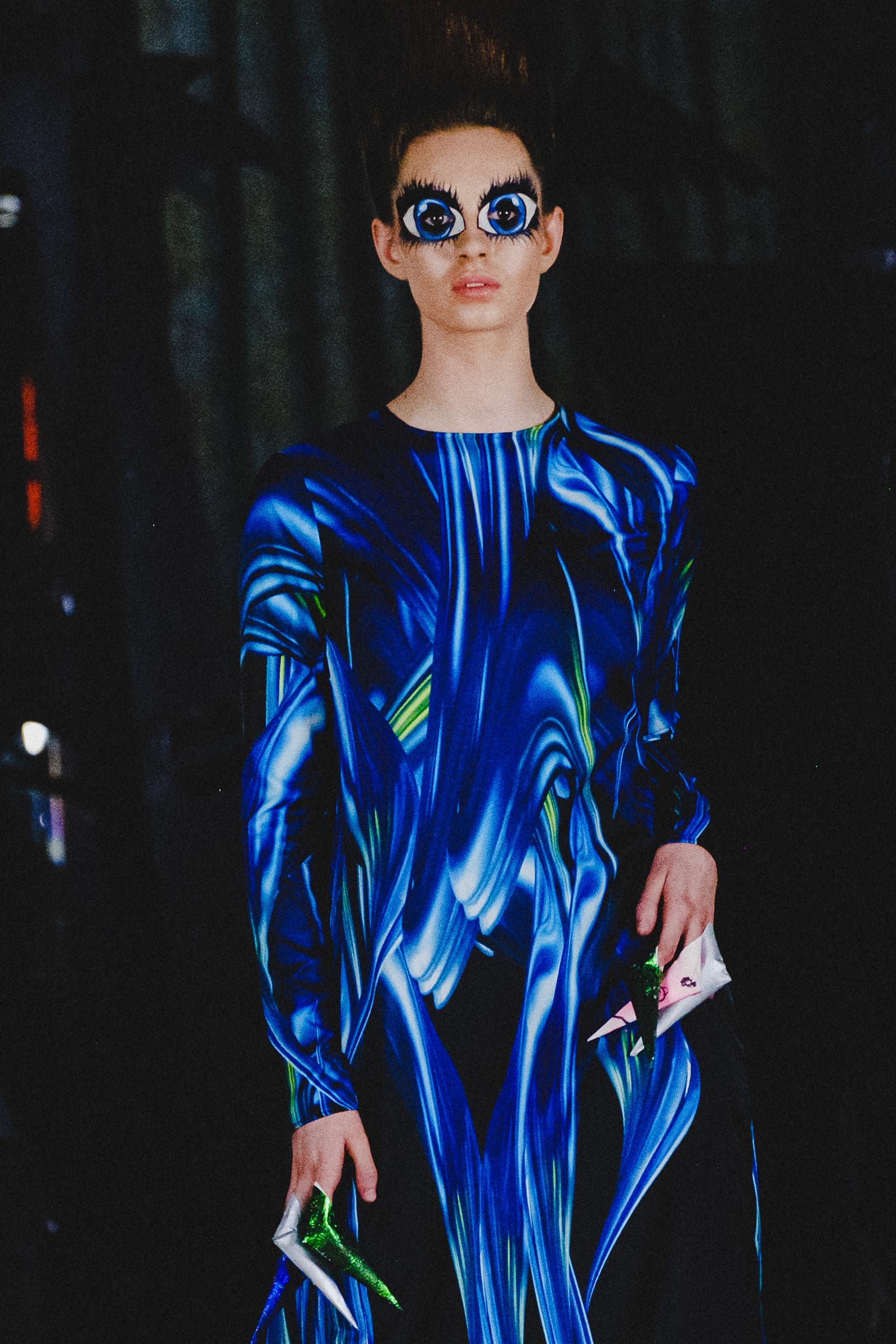 A person on a catwalk wearing a long-sleeved dress with a blue, black, and green swirling marble-like print. The person has exaggerated makeup around the eyes featuring large, illustrated blue irises with heavy black eyelashes. Their hair is styled in a tall, pointed fashion. They hold small, angular, metallic green and silver accessories in each hand against a dark, out-of-focus background.
