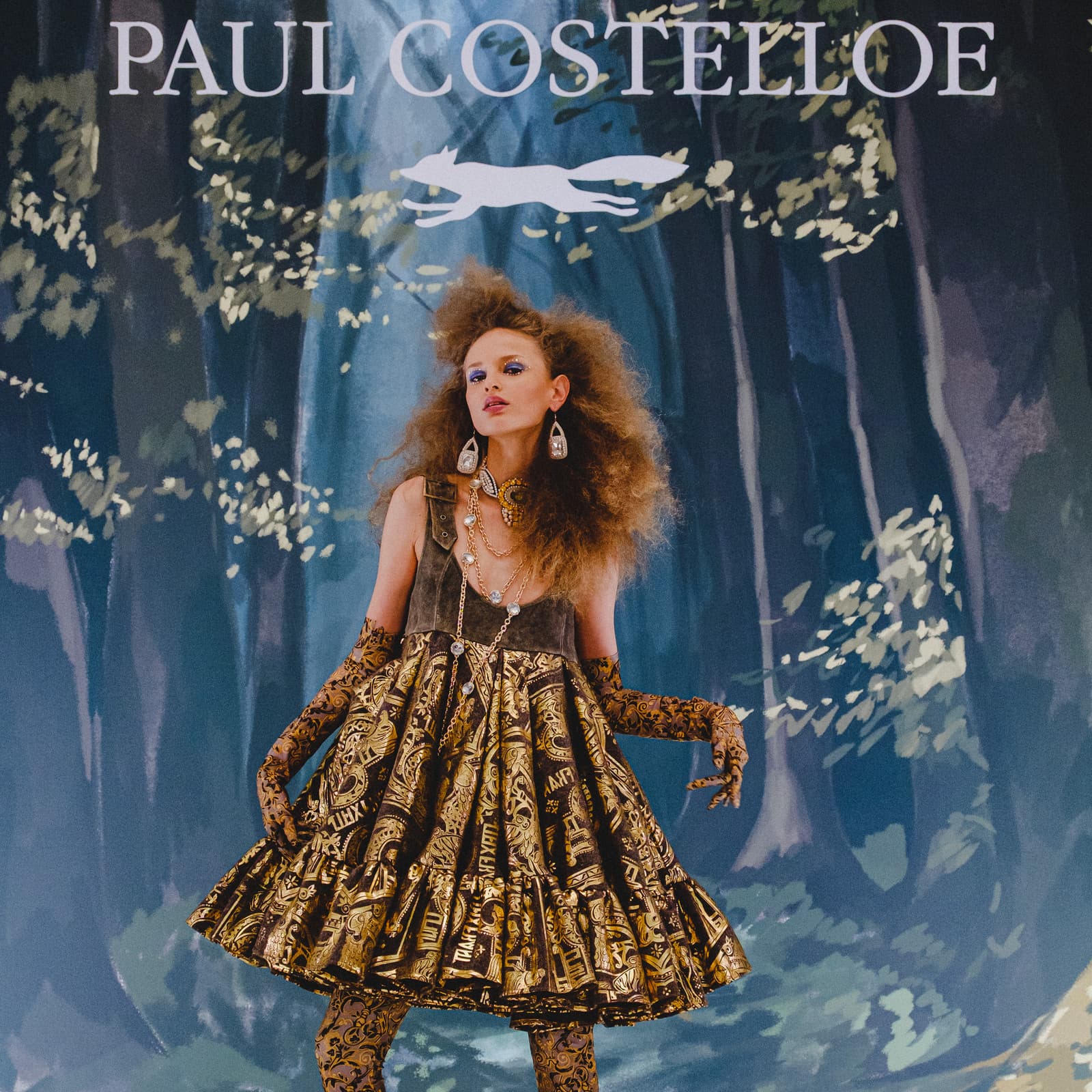 A model standing in front of a blue and green woodland-themed backdrop featuring a white fox icon and the text PAUL COSTELLOE. The model has voluminous, frizzy hair and wears a wide-strapped, textured grey-and-gold bodice dress with a gathered, patterned skirt featuring dark and metallic gold motifs. Underneath, the model wears matching semi-sheer, dark-patterned long gloves and hosiery. They are accessorized with multiple thin gold chains and large, pale earrings.
