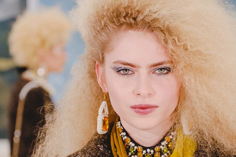A close-up portrait of a person with voluminous, crimped blonde hair facing the camera. They are wearing ornate pearl and amber jewelry around the neck and a large pearl-encrusted amber earring. Their eye makeup features sharp, winged eyeliner accented with small jewels. Another person with similar hair is partially visible and out of focus in the background.