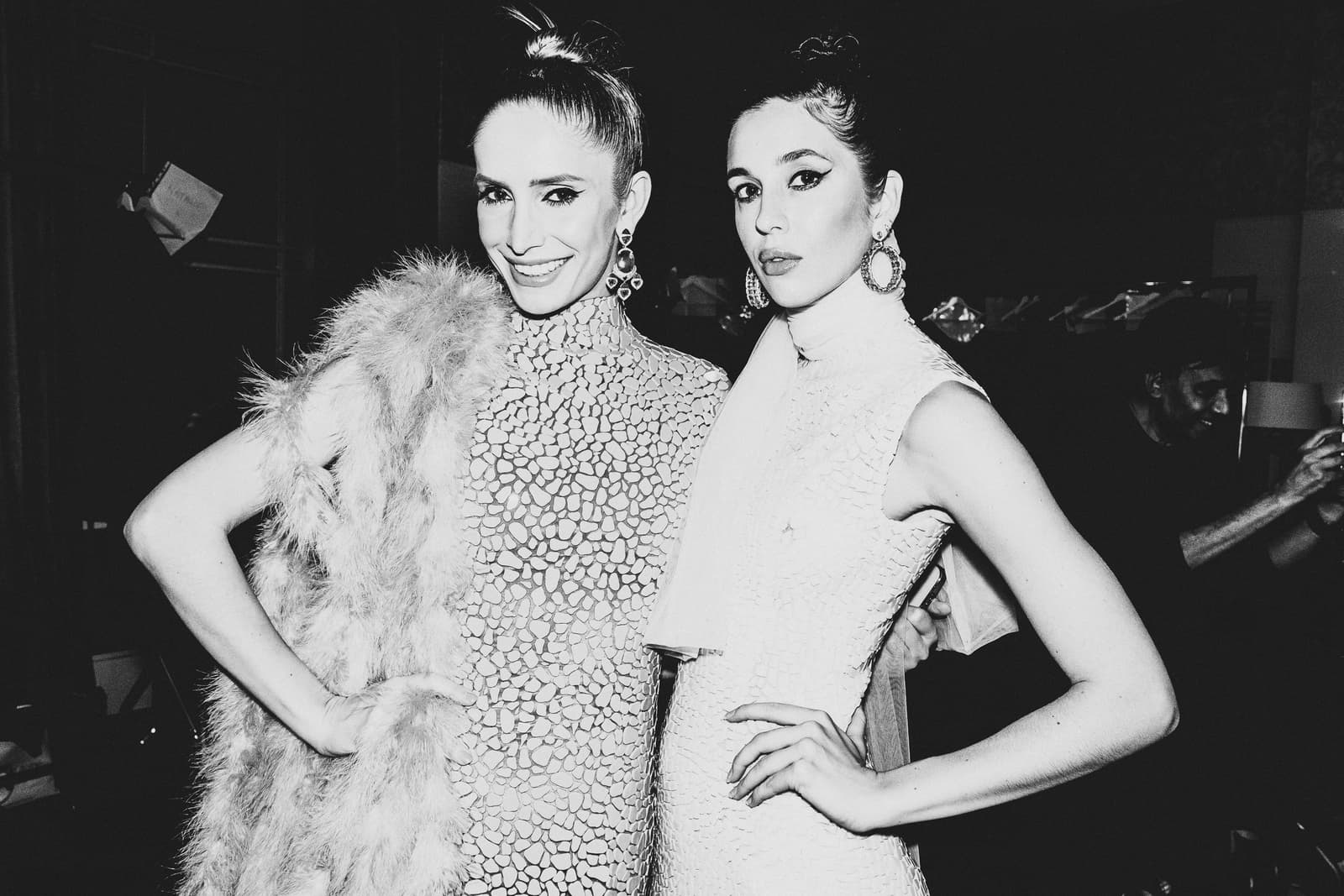 Two people standing side-by-side in a backstage setting, captured in black and white. Both individuals wear high-necked, textured sleeveless dresses and large, circular statement earrings. One person on the left has a feathered stole draped over one arm, while both have hair pulled back in tight updos. They are posed with hands on hips, facing the camera with neutral expressions.