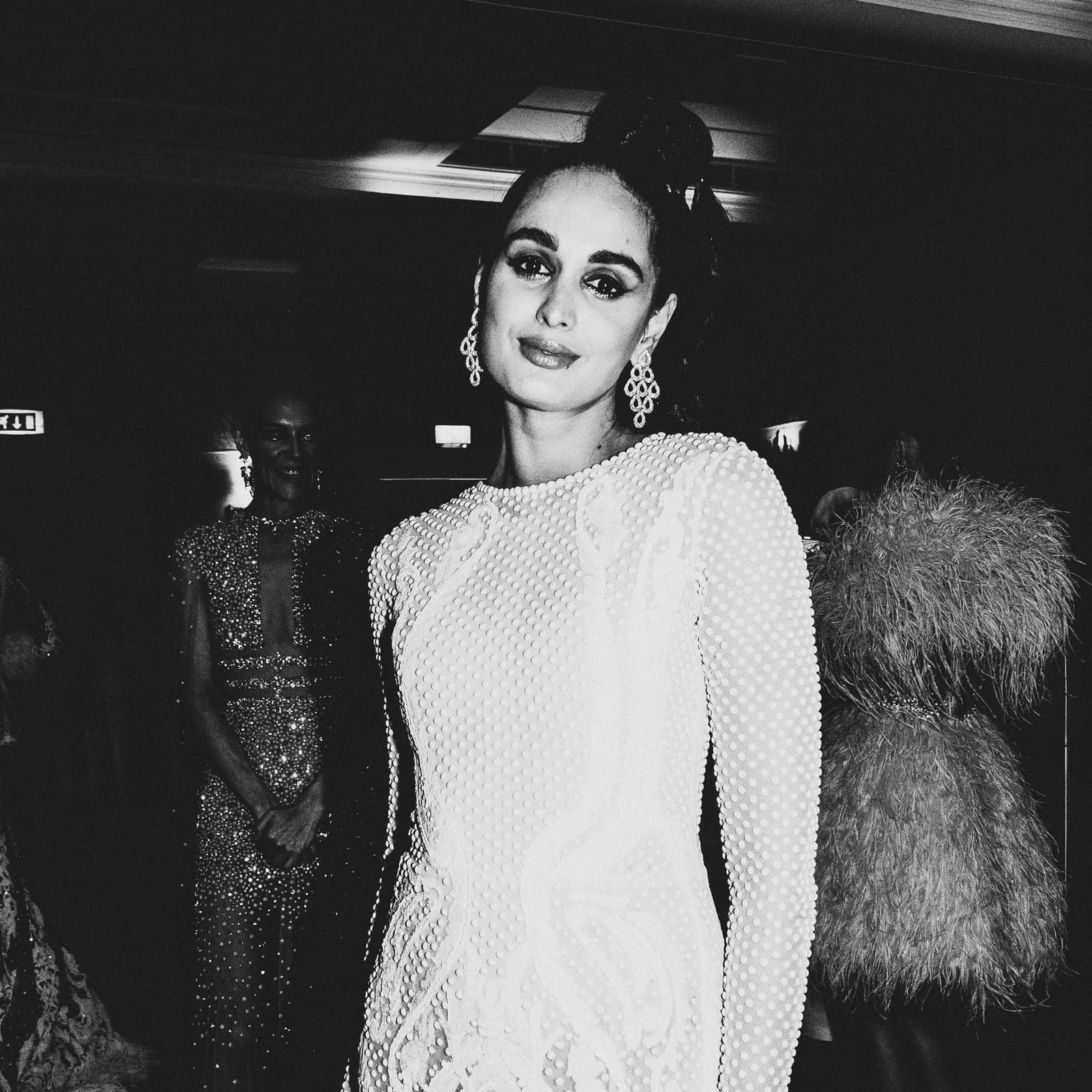 A medium shot of a model facing the camera in a dark indoor space, wearing a white, textured, long-sleeved gown with a repeating beaded pattern. She has a high bun and wears large, ornate earrings. In the dimly lit background, another person in a sequined dress and a third person in a feathery garment are visible.