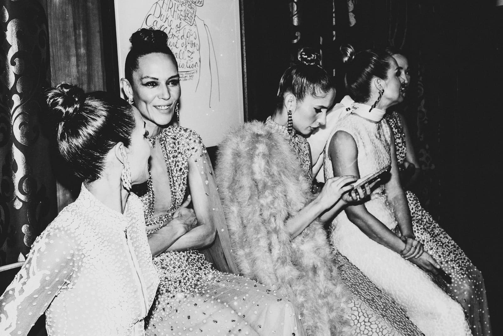 A black-and-white medium shot of four individuals sitting in a row backstage at a fashion show. The people wear heavily embellished gowns with beadwork and fringe, with their hair pulled back in high buns. They are illuminated by bright, directional lighting that creates high contrast against the dark background, and one person looks down at a mobile phone.